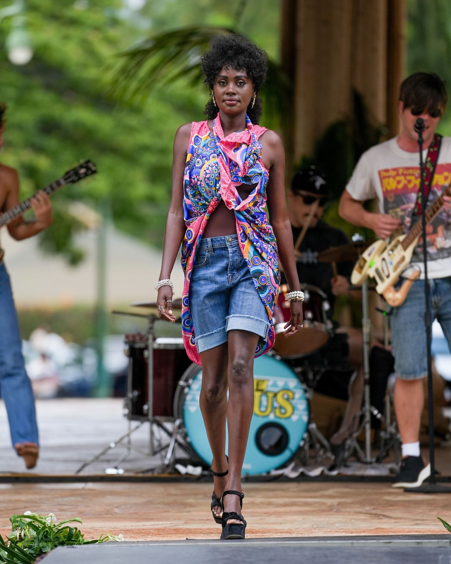 A performer in denim and colorful attire dances energetically on an outdoor stage during a summer concert.