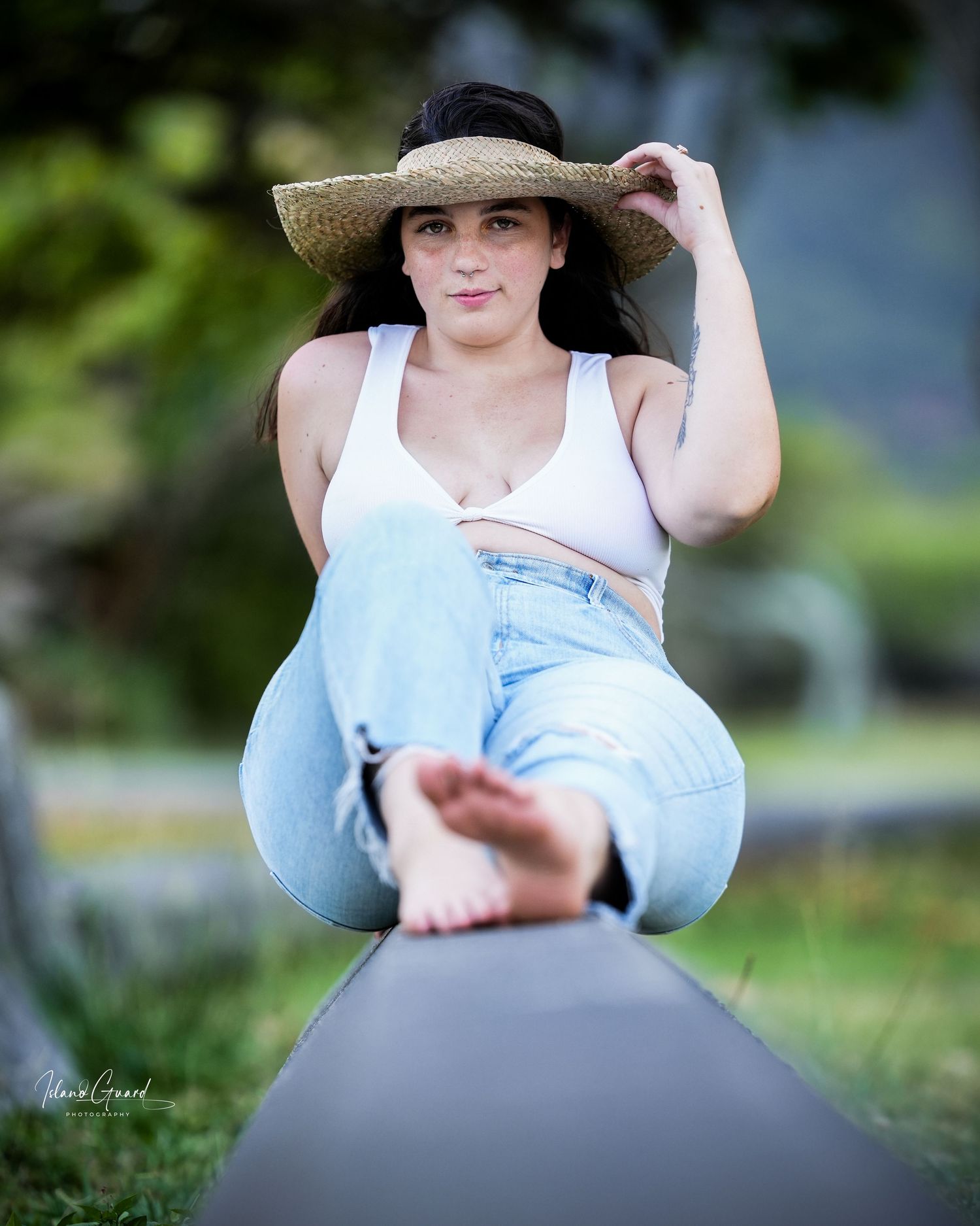 A barefoot model reclines on a log wearing a straw hat, white tank top and jeans in an outdoor garden setting.