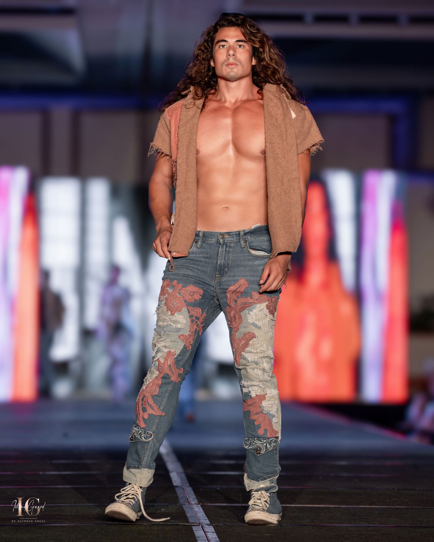 Model showcasing distressed denim jeans and tan jacket on fashion runway with dramatic lighting.