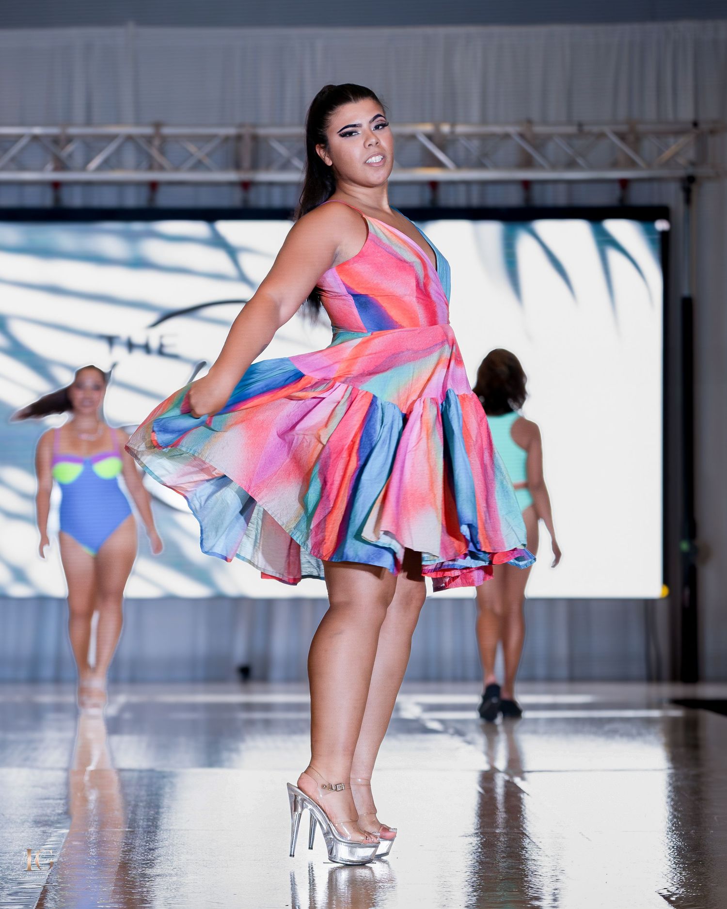 Models strut in colorful flowing dresses on a mirrored catwalk during a fashion presentation.