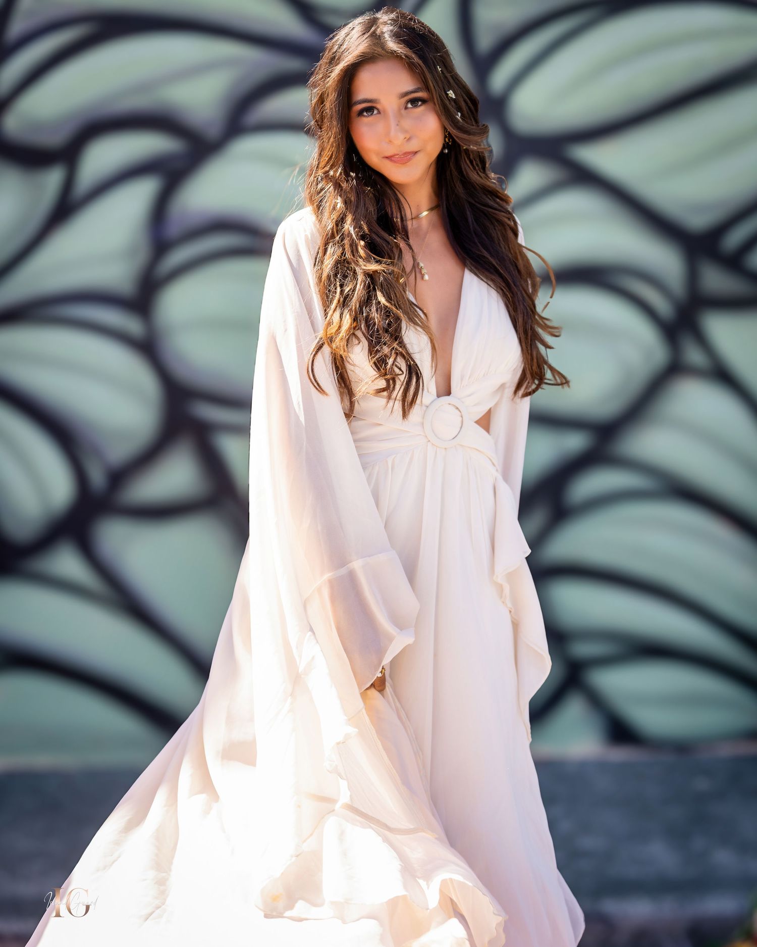 Model in white flowing outfit poses elegantly against urban art wall background.
