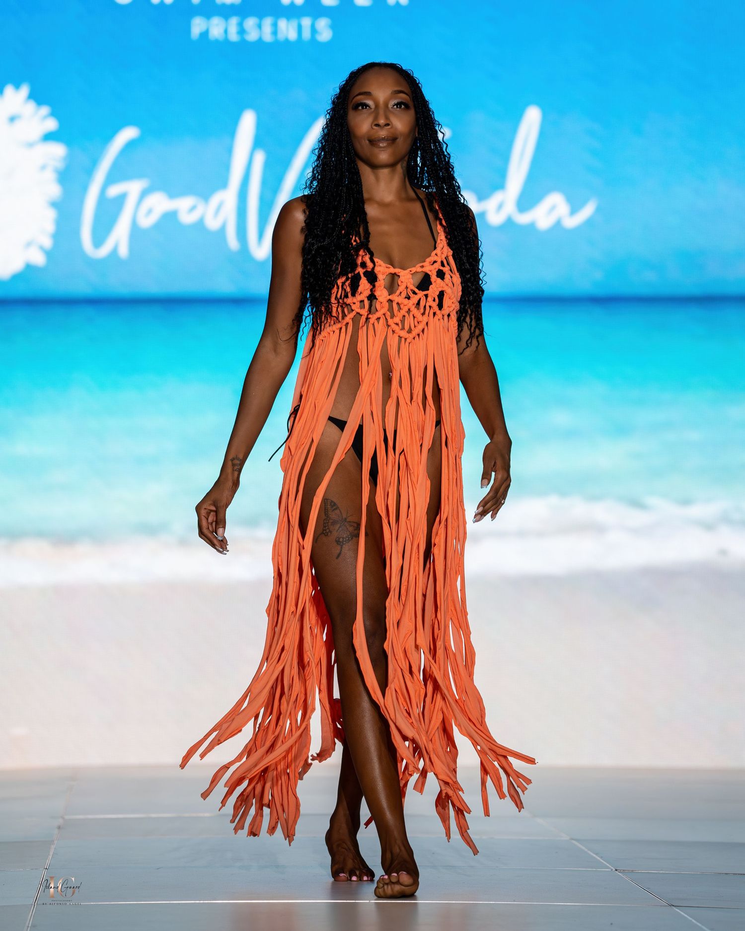 Coral fringe swimwear cover-up dress showcased on runway with tropical beach backdrop.
