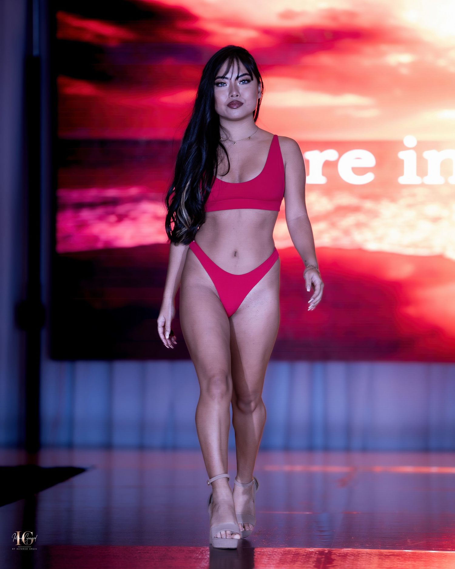 Model walks runway in red bikini during fashion show with dramatic red lighting and backdrop.
