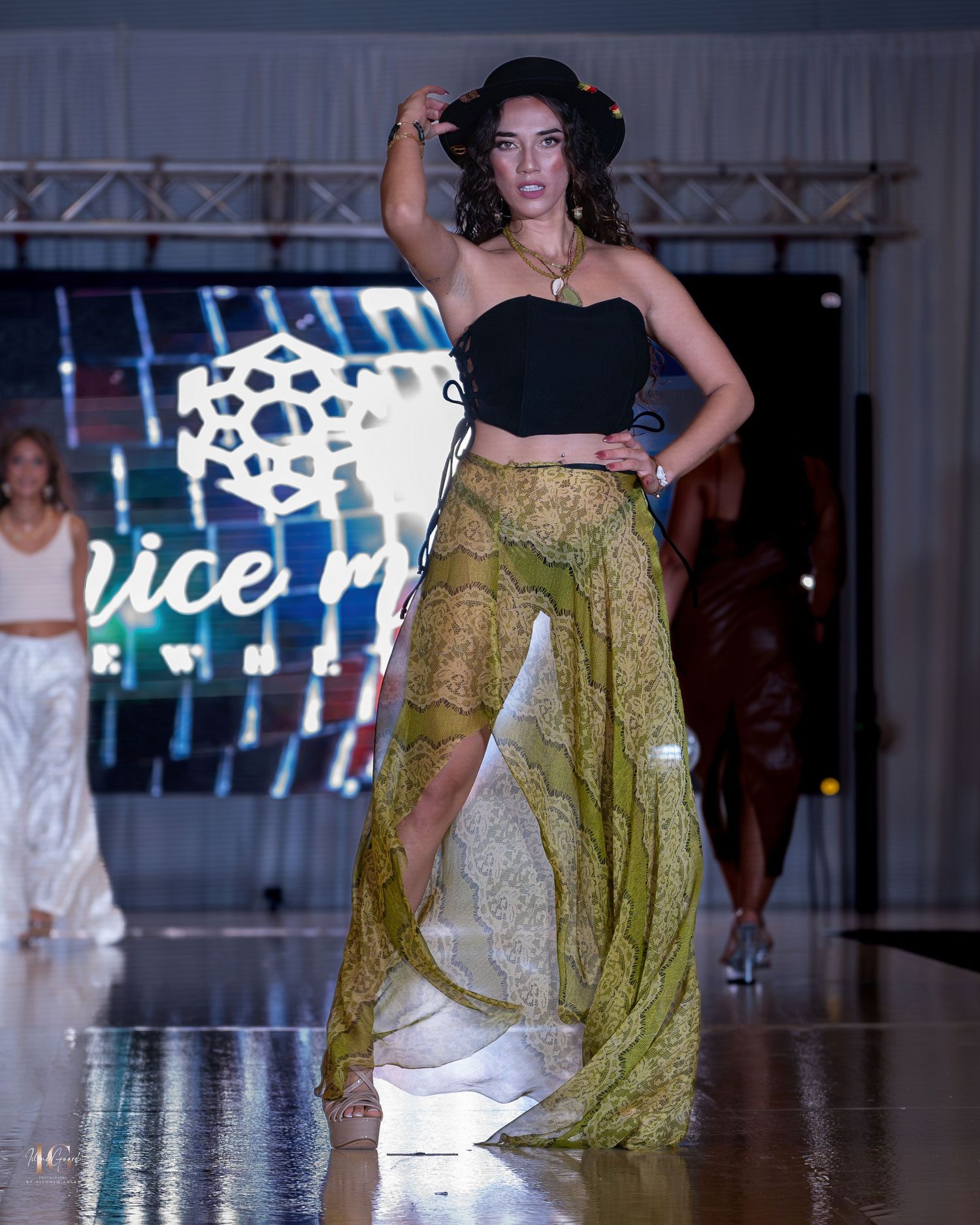 Fashion model walks runway in black crop top and olive green flowing skirt at fashion show.