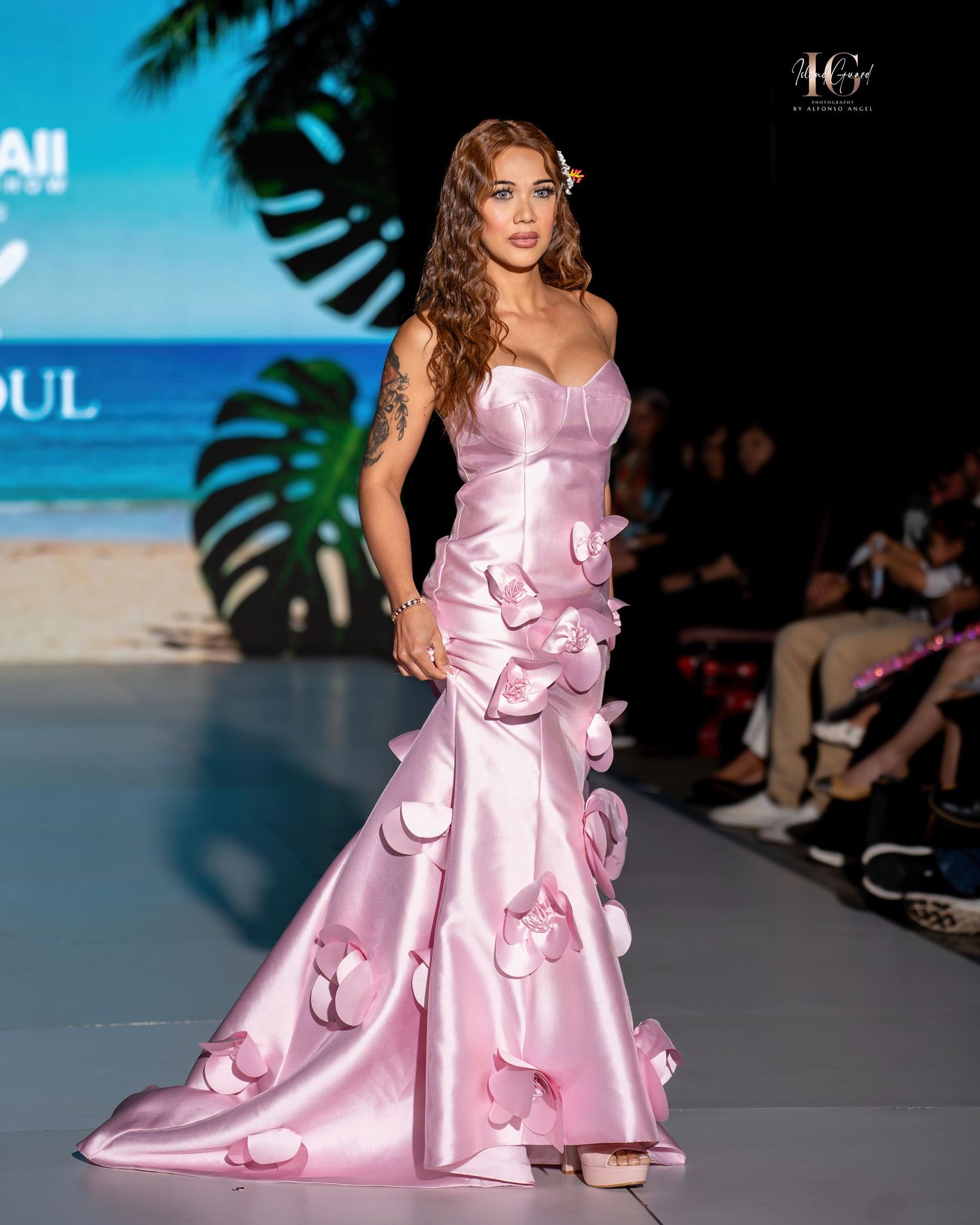 A model walks the runway in a pink satin mermaid gown with floral appliques at a tropical-themed fashion show.