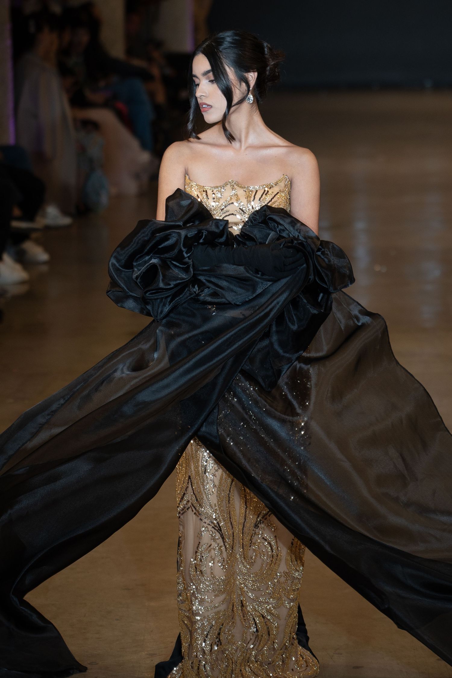 Fashion model wearing dramatic black and gold evening gown with flowing cape detail on runway.