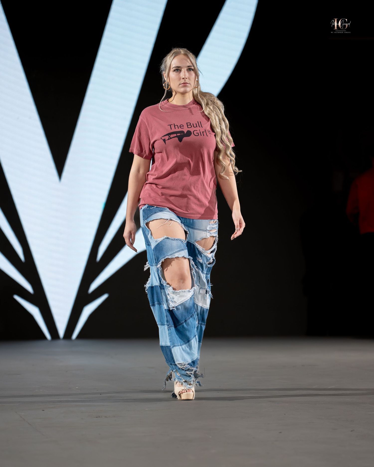 Model walks runway in distressed denim jeans and burgundy t-shirt during fashion show.