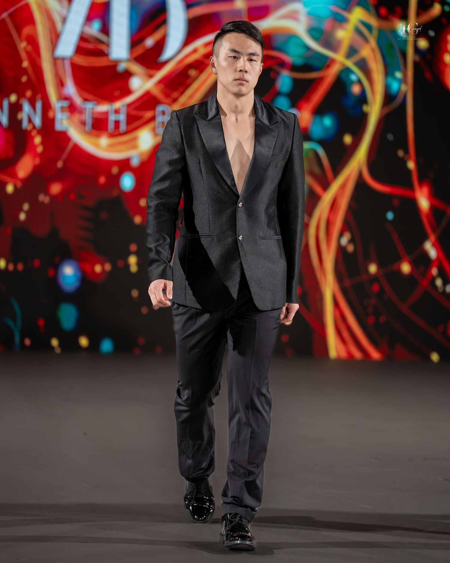 A performer in a black suit strikes dynamic poses during a stage performance with colorful lights in the background.