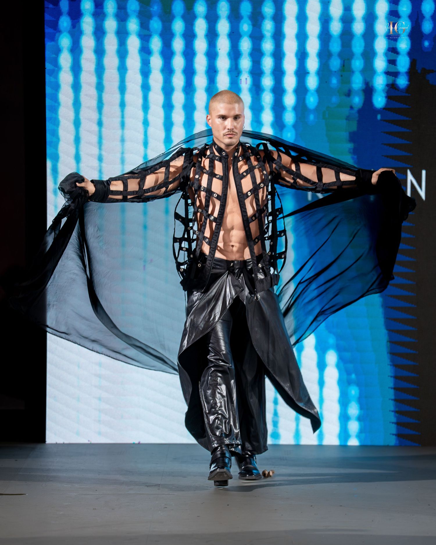 Model walks runway in dramatic black flowing coat with billowing panels against blue digital backdrop during fashion show.