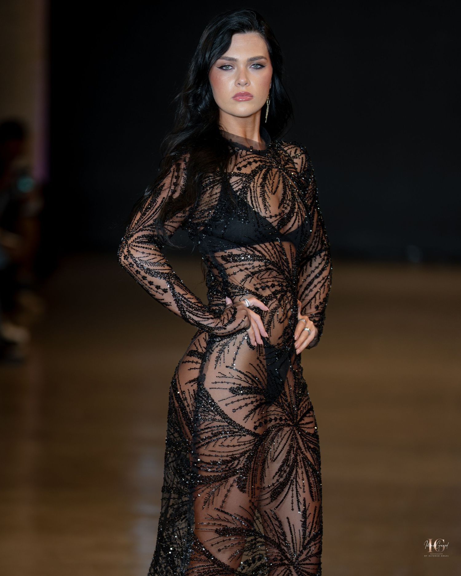 Fashion model walks runway in sheer black lace evening gown with long sleeves against dark background.
