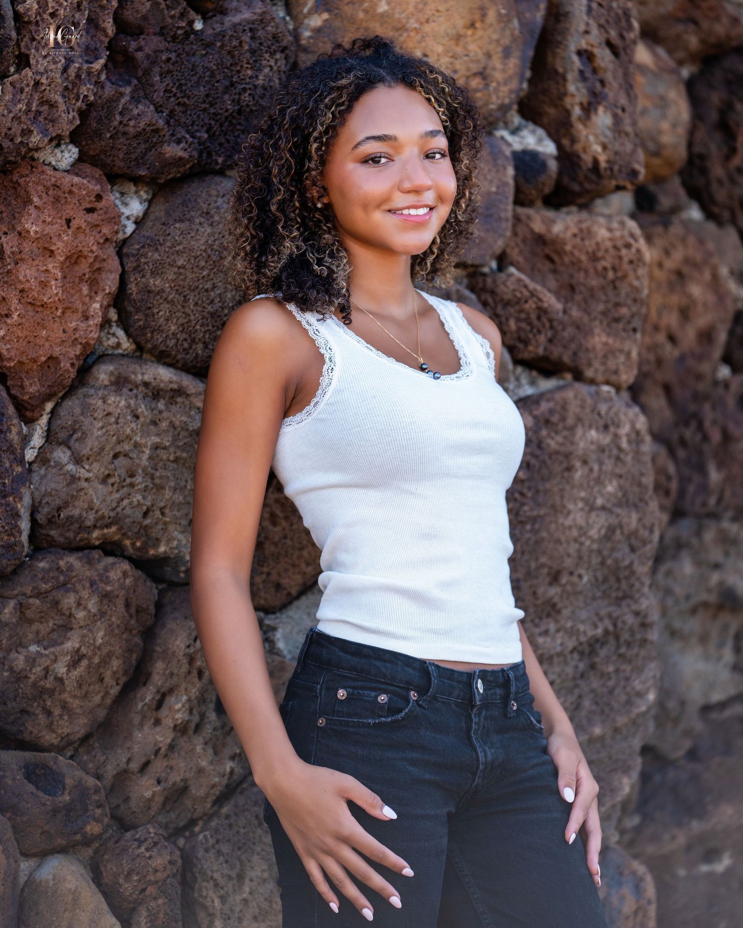 Young woman in white tank top and jeans smiles while posing against rustic stone wall outdoors.