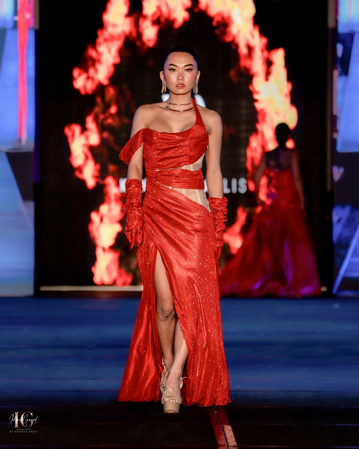 One-shoulder red evening gown with high slit and dramatic backdrop of flames on fashion runway.