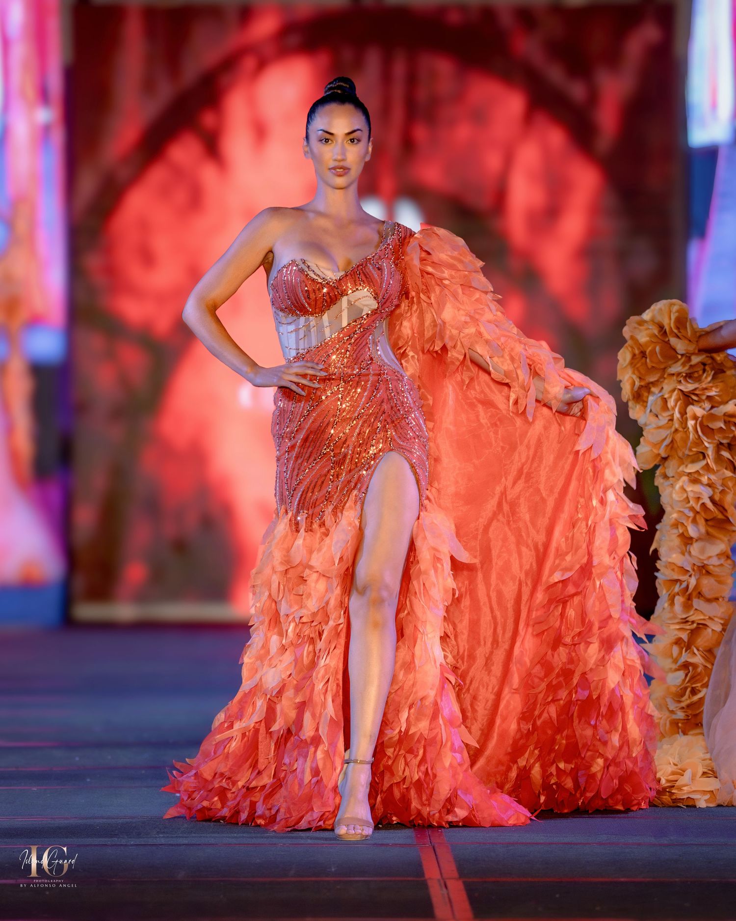 Flowing coral orange ruffled gown with dramatic train displayed on illuminated catwalk.