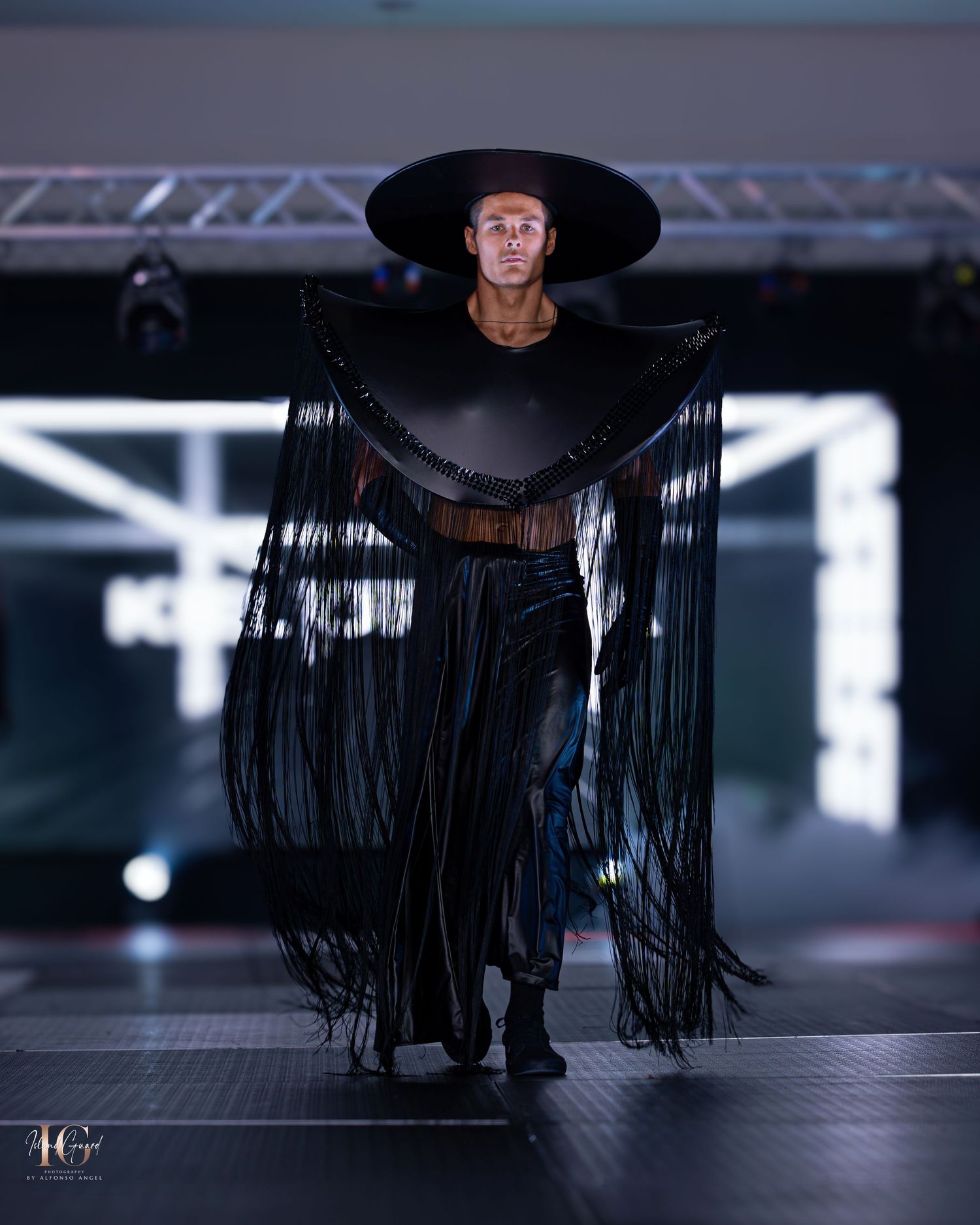 Dramatic black wide-brim hat with flowing fringe details displayed on darkened catwalk.