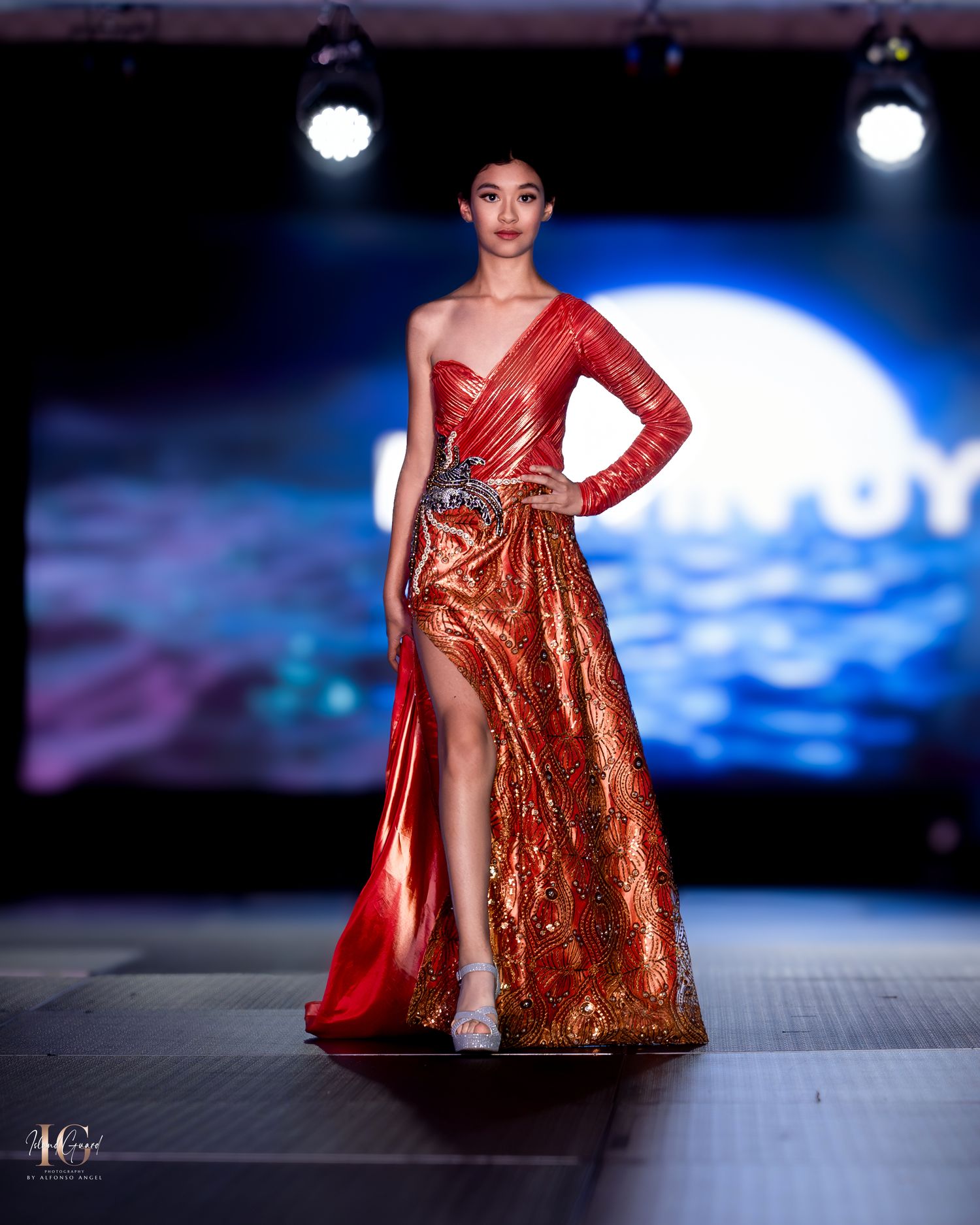 Model showcases red sequined evening gown with dramatic slit on illuminated runway.