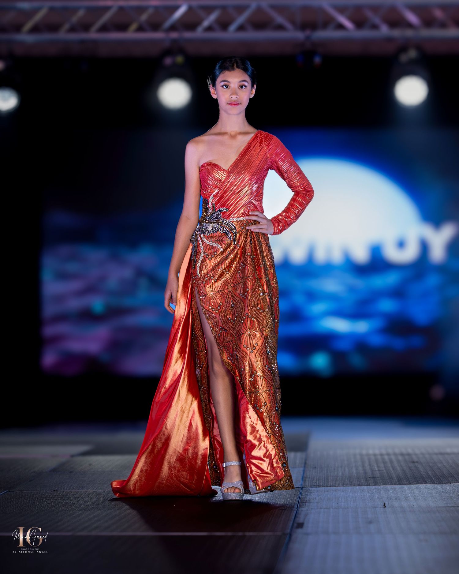 Fashion model walks runway in stunning red and gold evening gown with one-shoulder design and thigh-high slit.