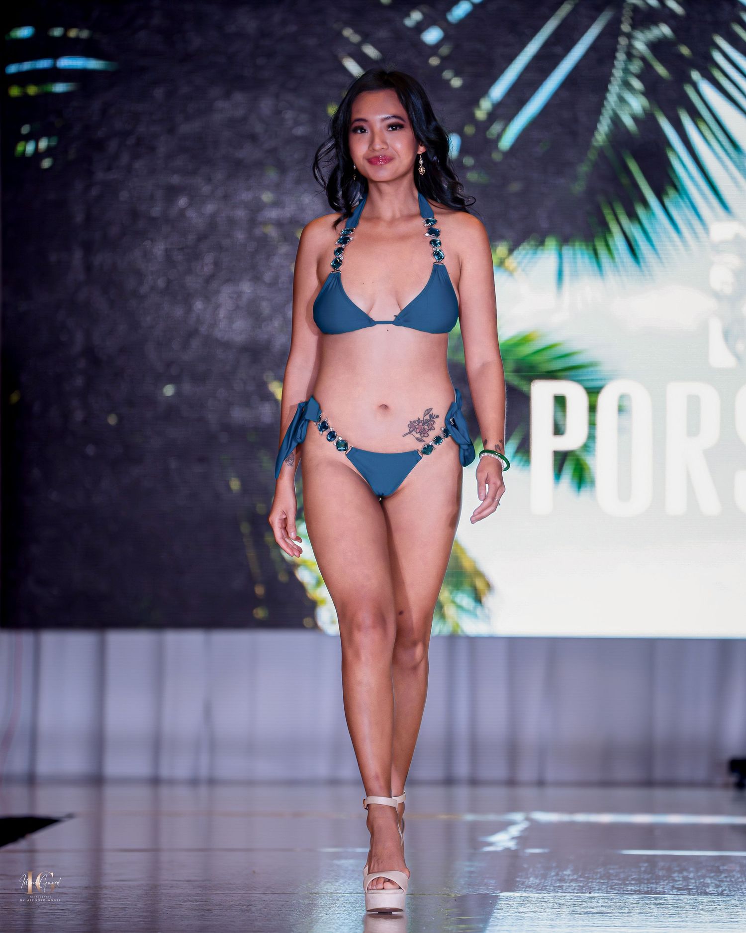 Models presenting blue swimwear collection on fashion runway with tropical background.