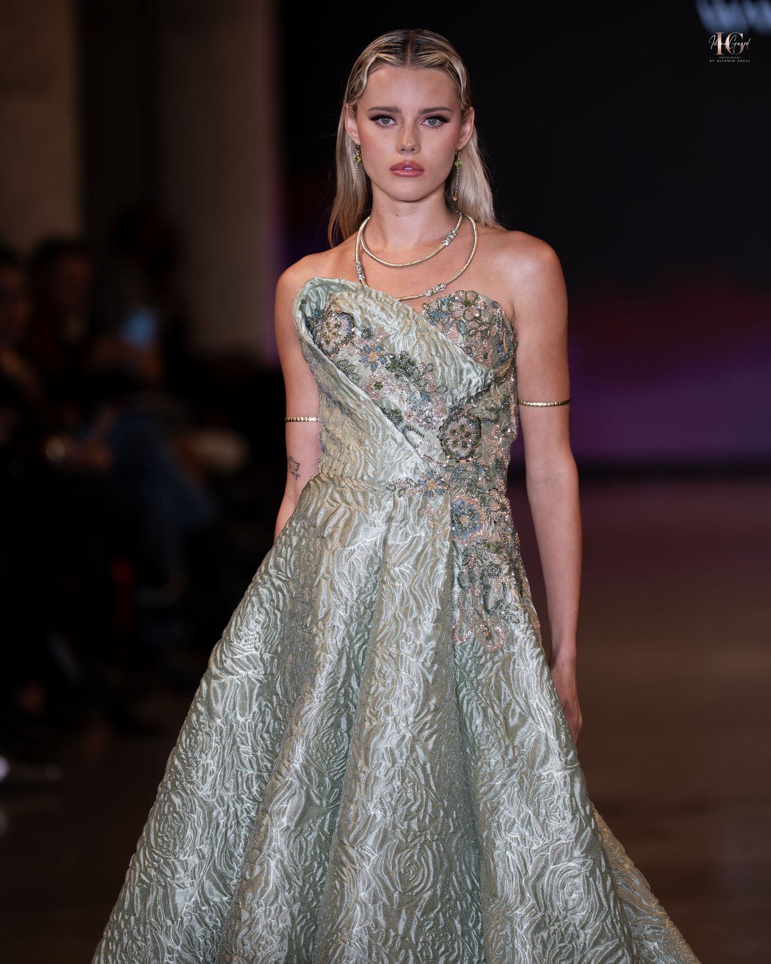 Model walks runway in metallic silver brocade babydoll dress during fashion show.