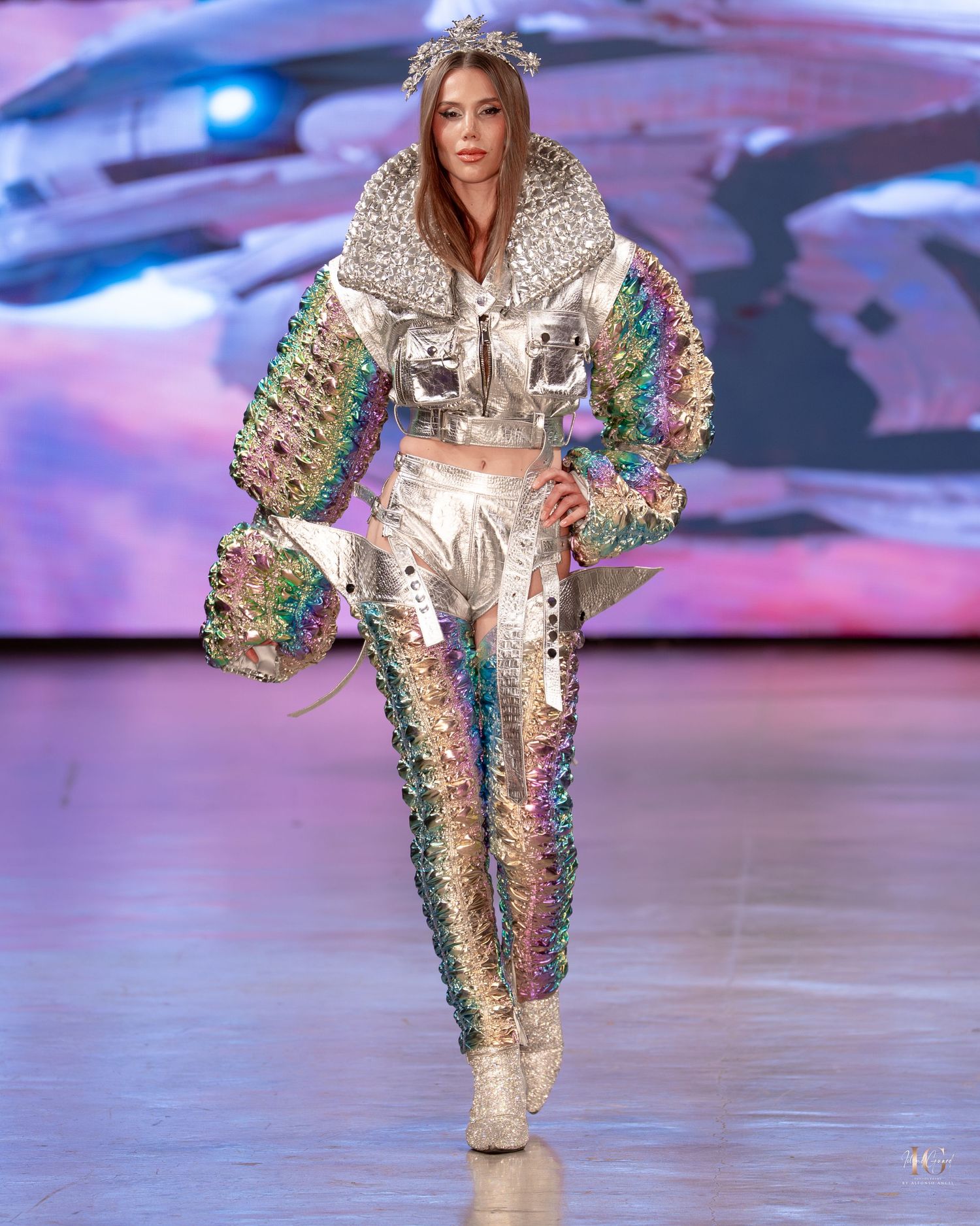A model walks the runway in a metallic silver jumpsuit with rainbow sequined sleeves at a fashion show.
