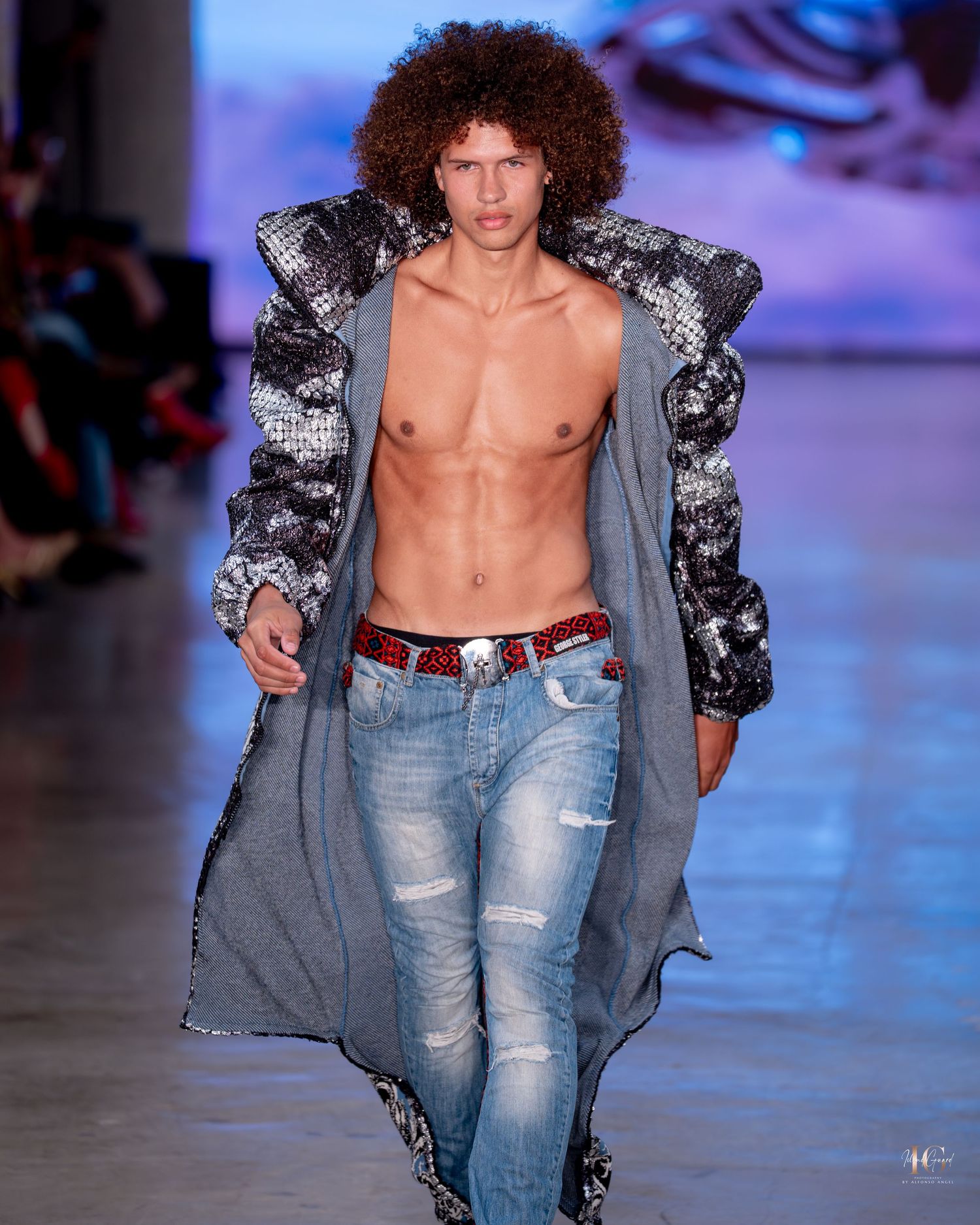 Model walks runway wearing distressed denim jeans and open sequined jacket showing bare chest at fashion show.