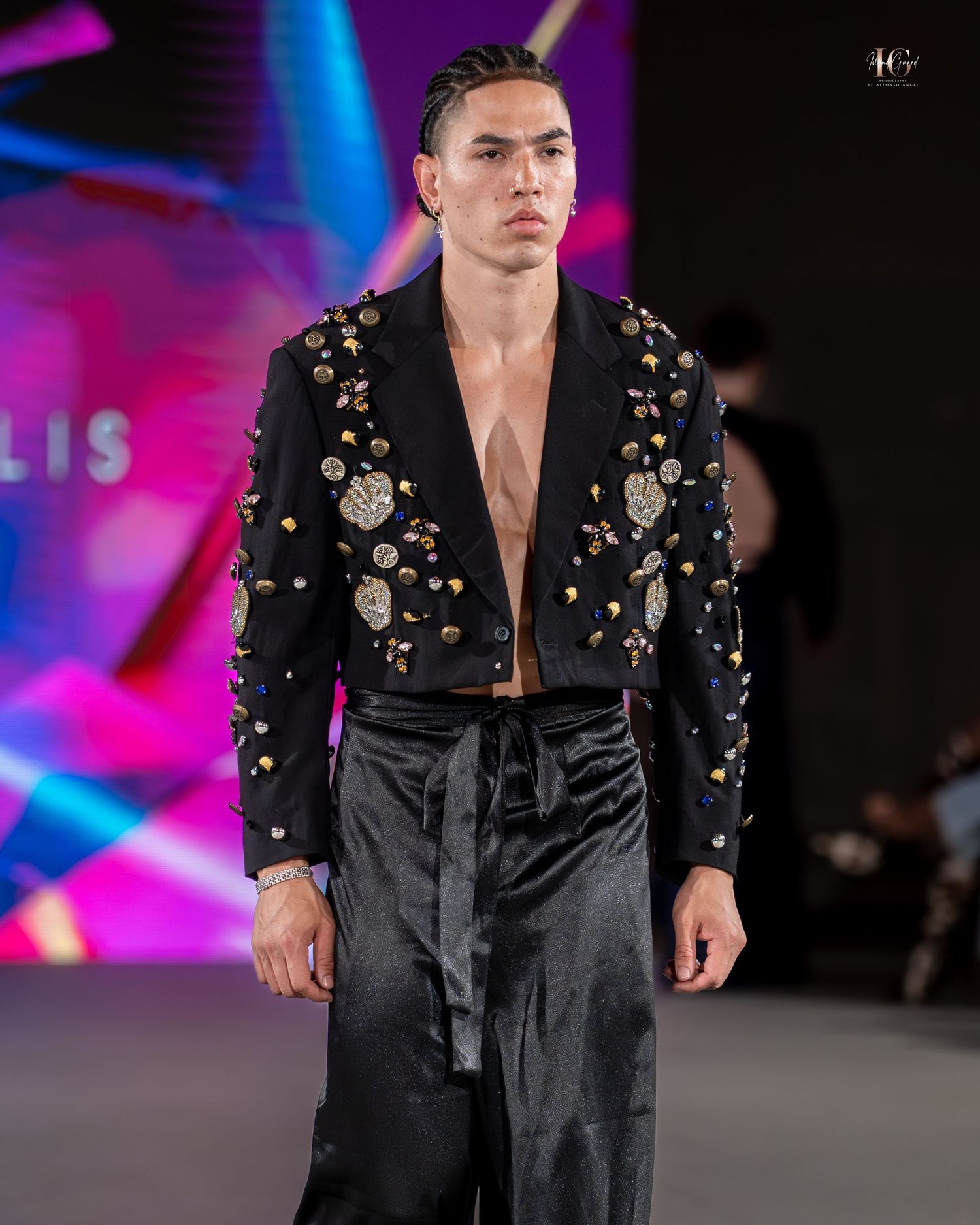 Model walks runway in black embellished jacket with studded shoulders and deep v-neck during fashion show.