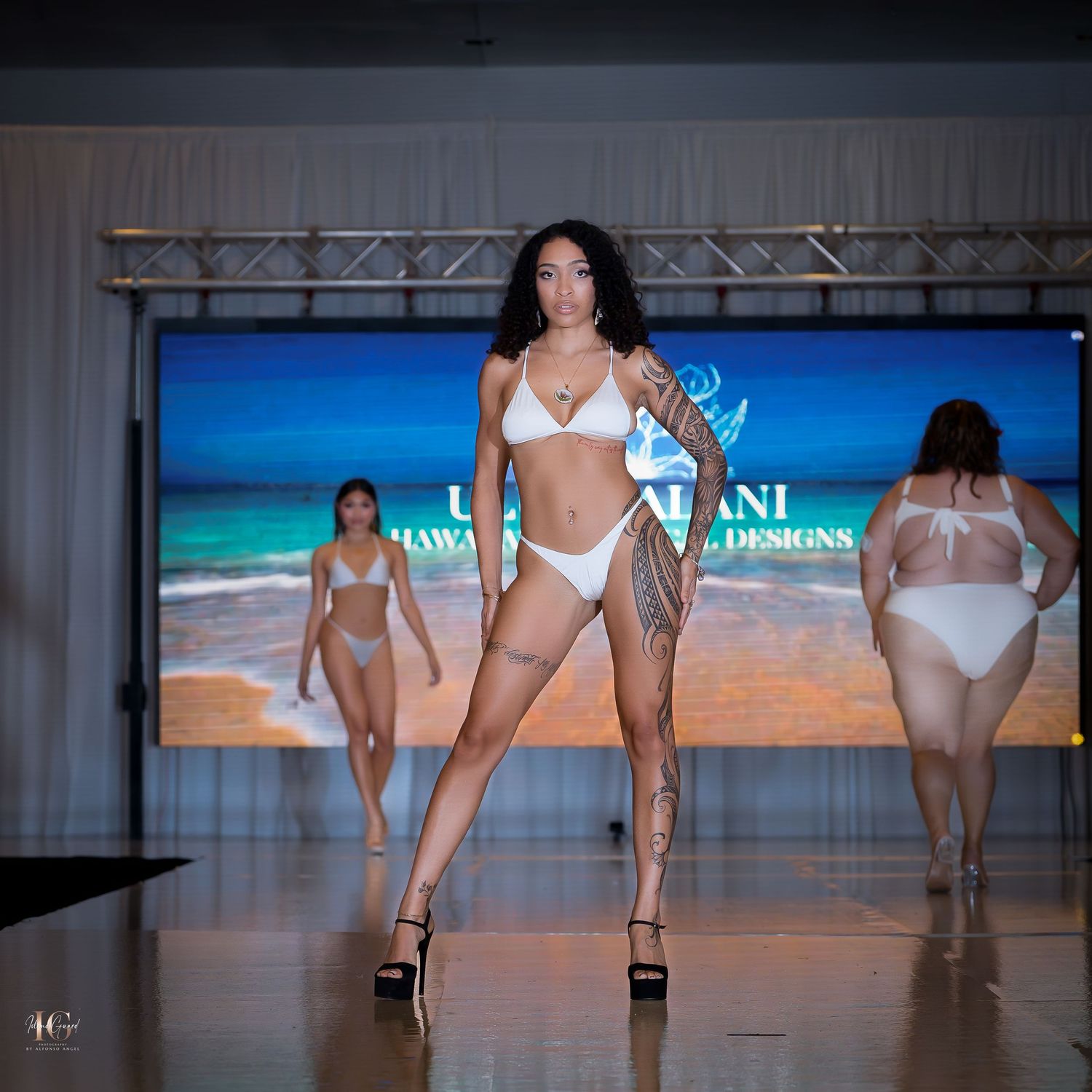 Models walk the runway in white swimwear during a fashion show with a tropical beach backdrop display.