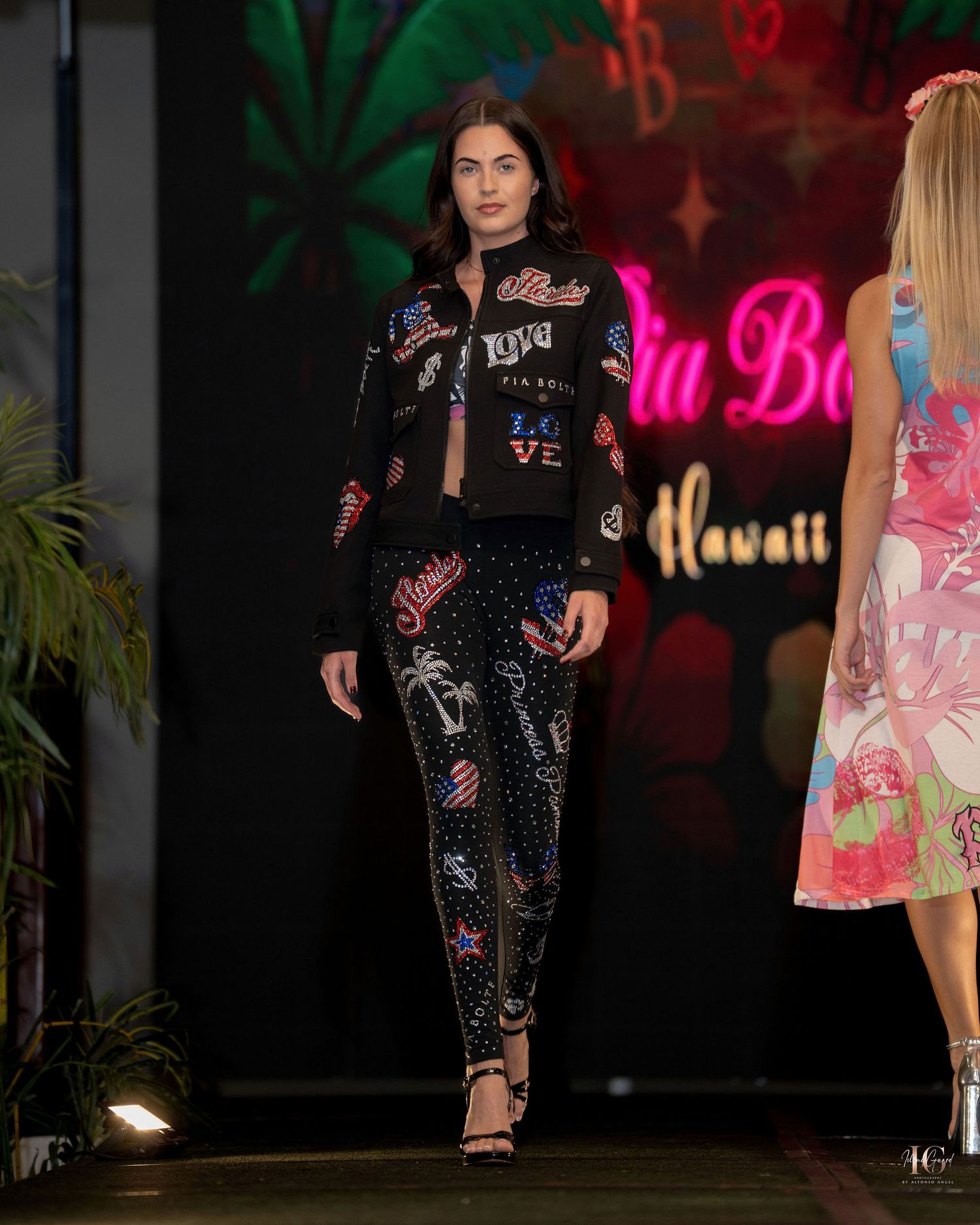 Models walk the runway wearing black embellished evening wear with tropical motifs at a fashion show.