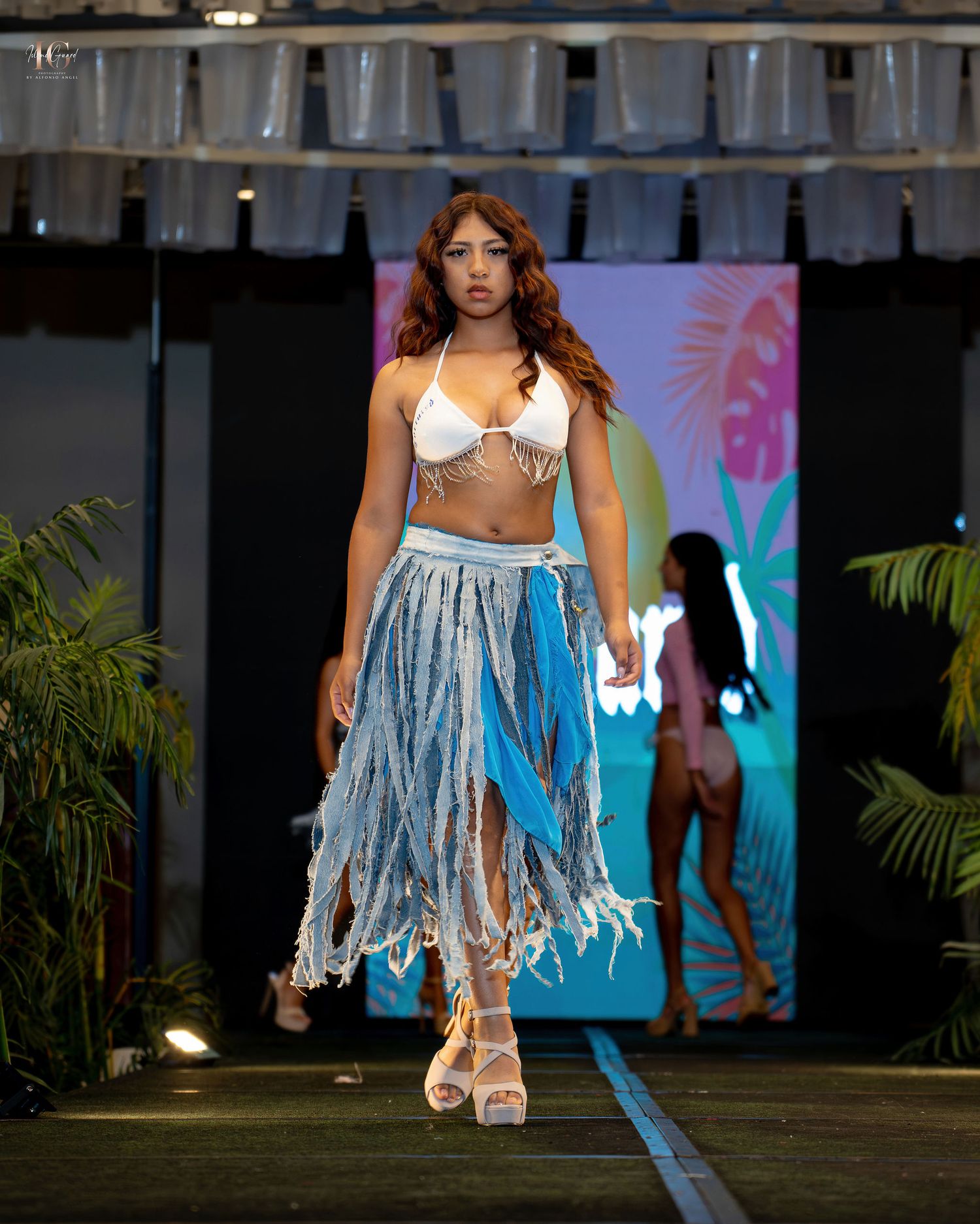 Model walks runway in white bikini top and tattered denim skirt with fringe details during tropical-themed fashion show.