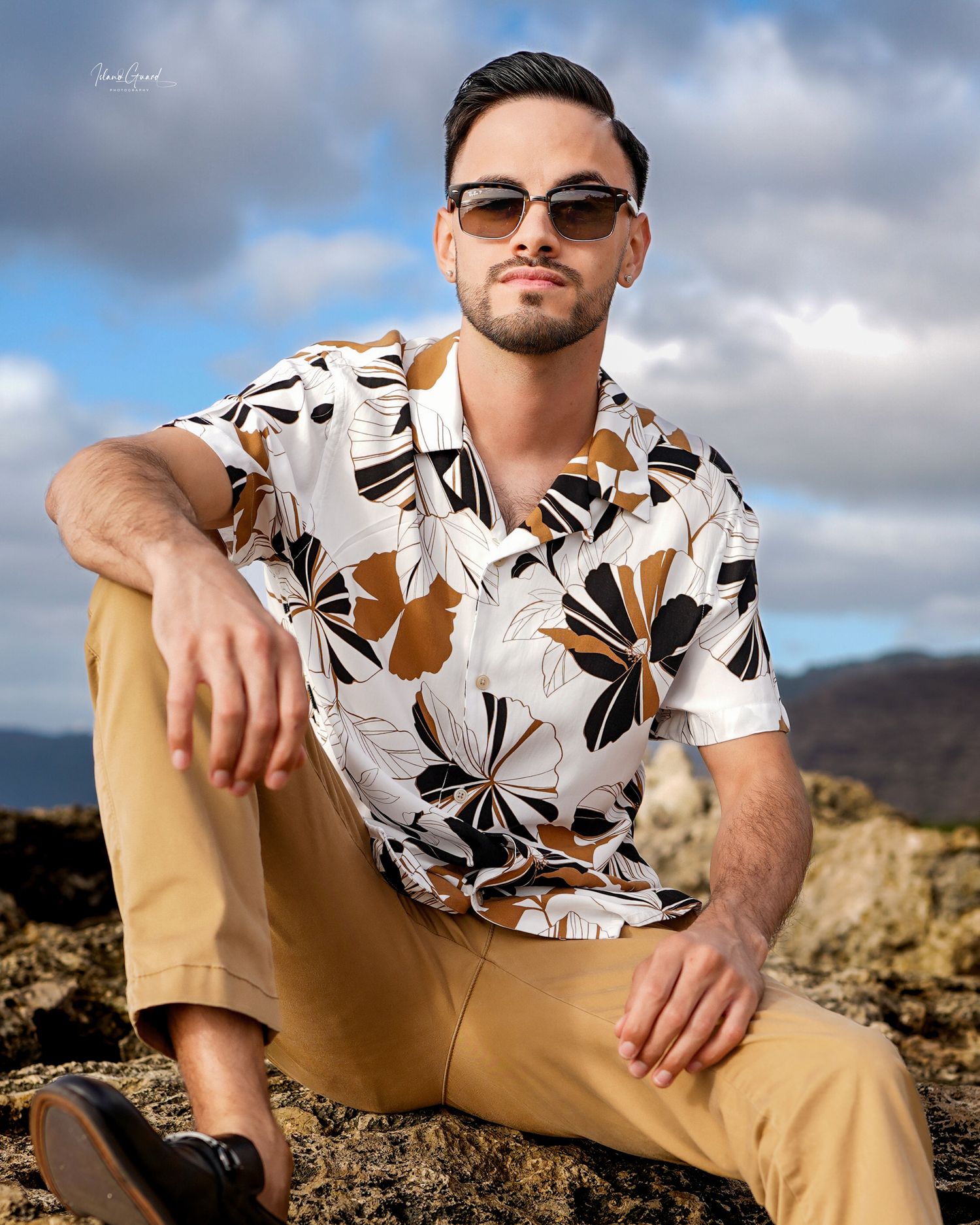 A person in a floral print shirt and khaki pants poses confidently on rocky terrain against a cloudy mountain backdrop.