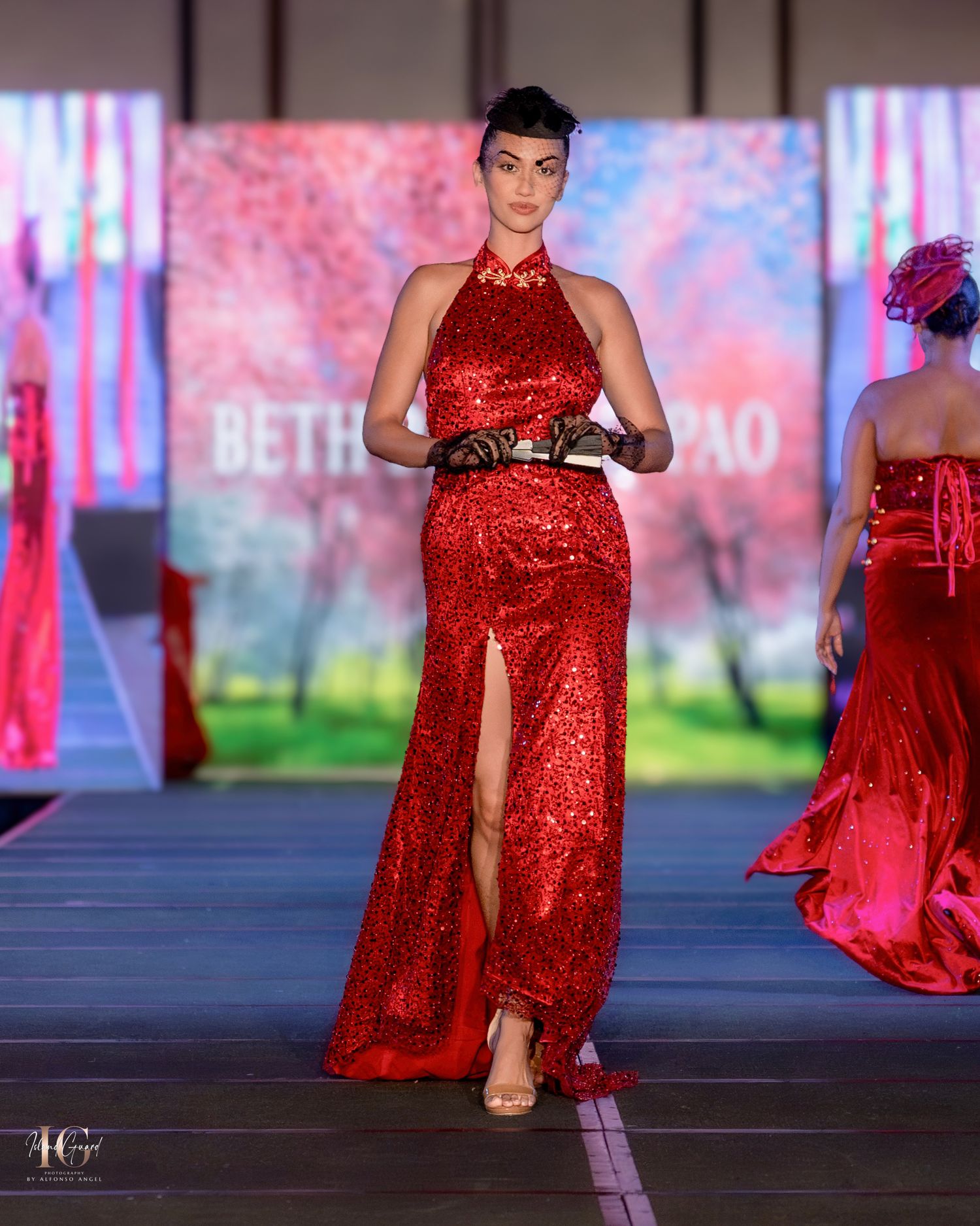 Model showcases sleek red halter gown with thigh-high slit on fashion runway with colorful backdrop.