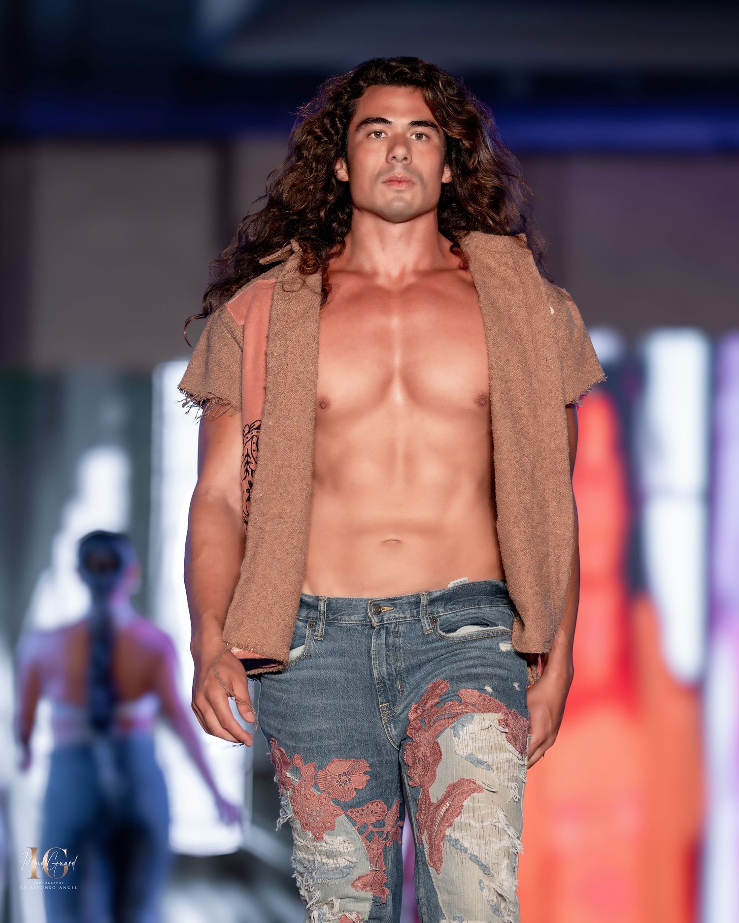 Model wearing tan suede jacket and distressed jeans walks dramatic runway with blue lighting.
