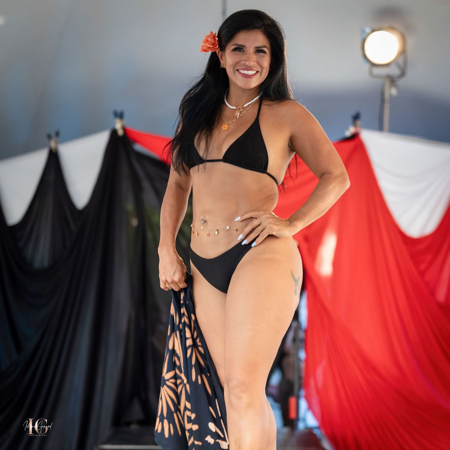 Model walking runway in black swimwear against red and black stage backdrop.