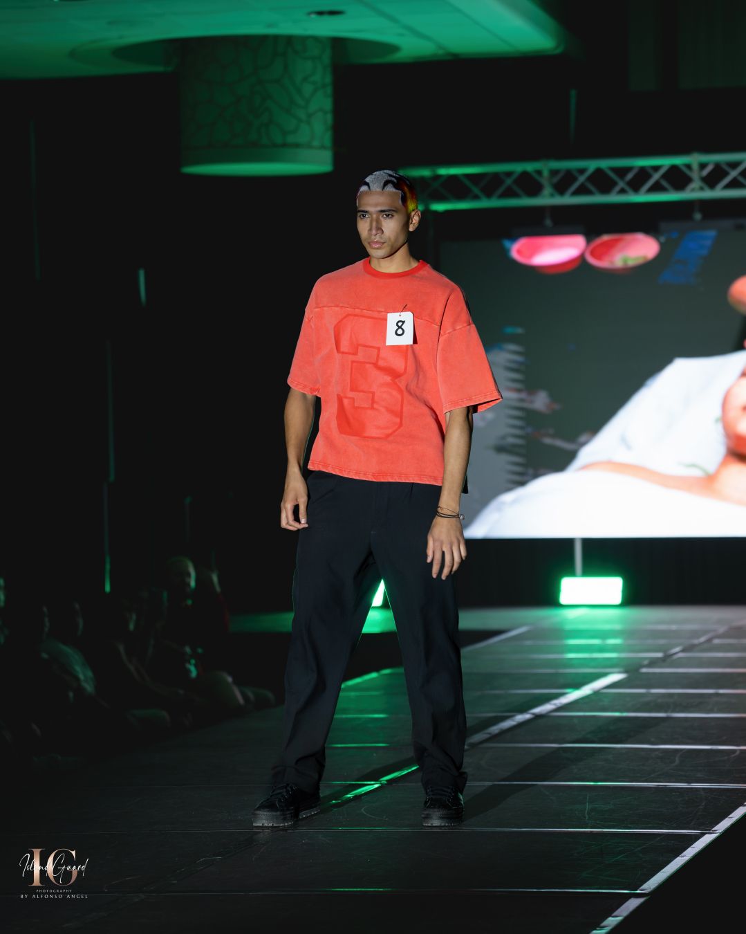 Model walks fashion runway in dark venue with green stage lighting.