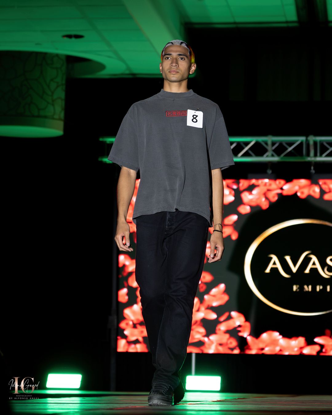 Model presents oversized grey t-shirt with black pants on dimly lit runway stage.