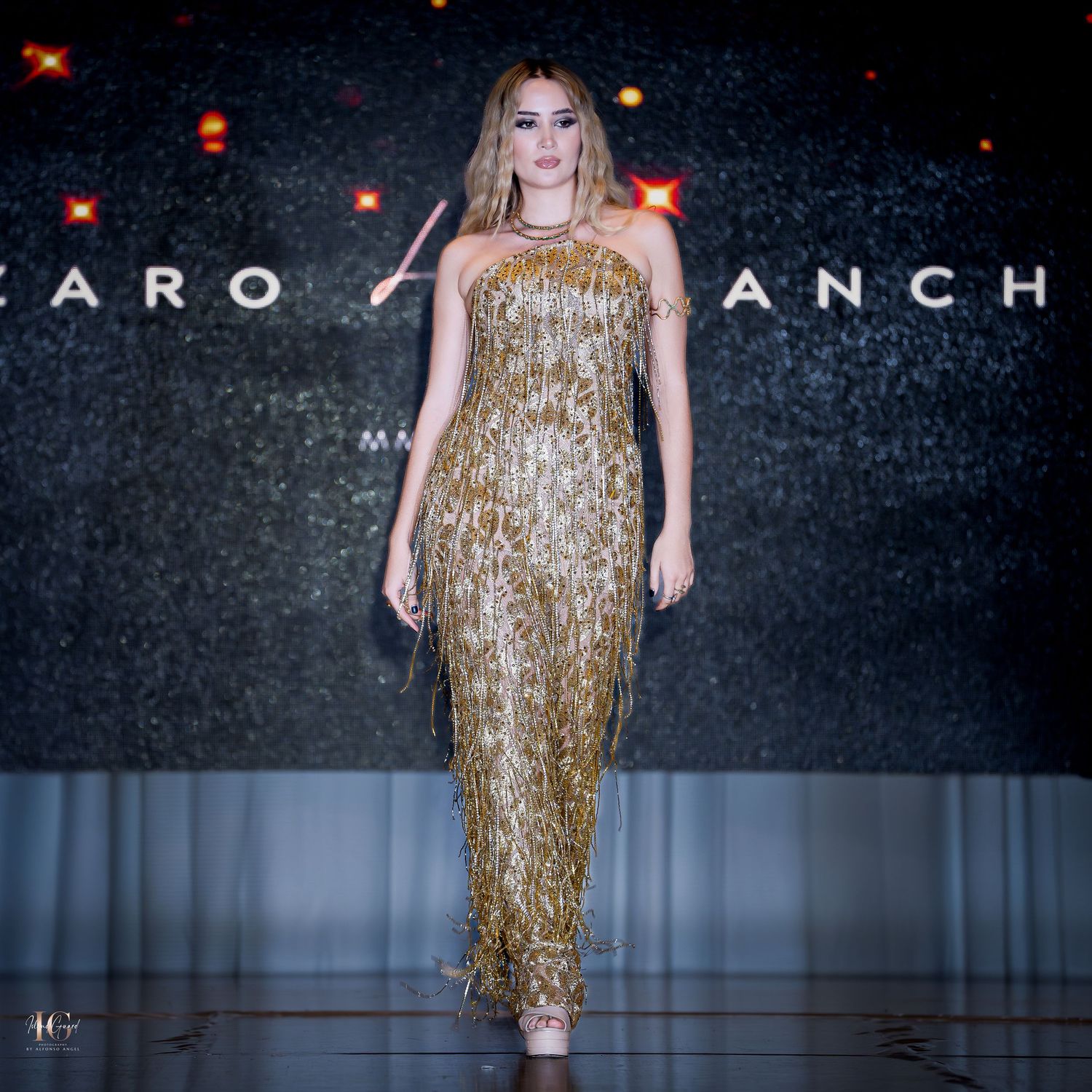 Stunning gold fringe gown displayed on fashion runway with dark backdrop.