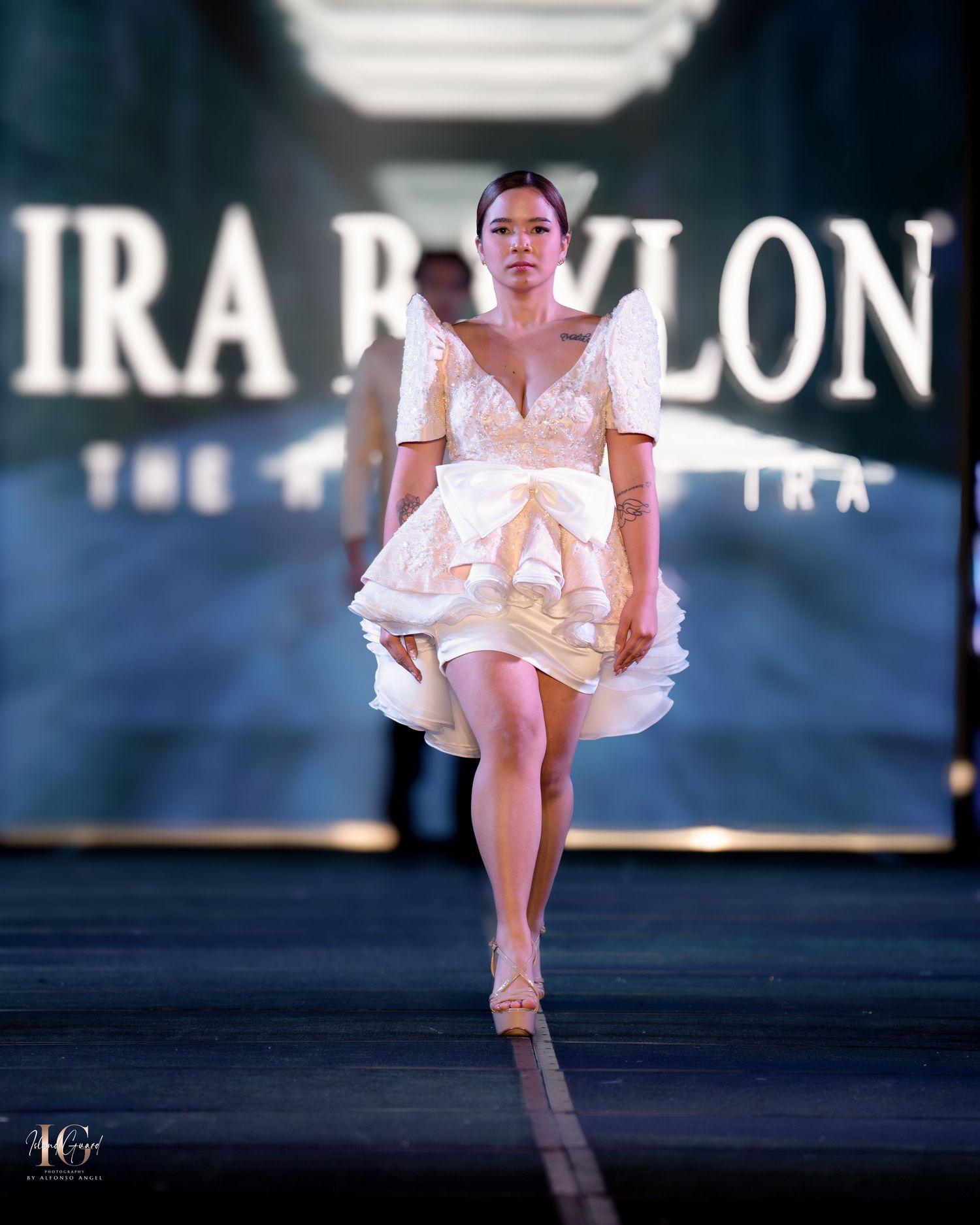Model in white ruffled dress performs dynamic runway walk against illuminated backdrop.