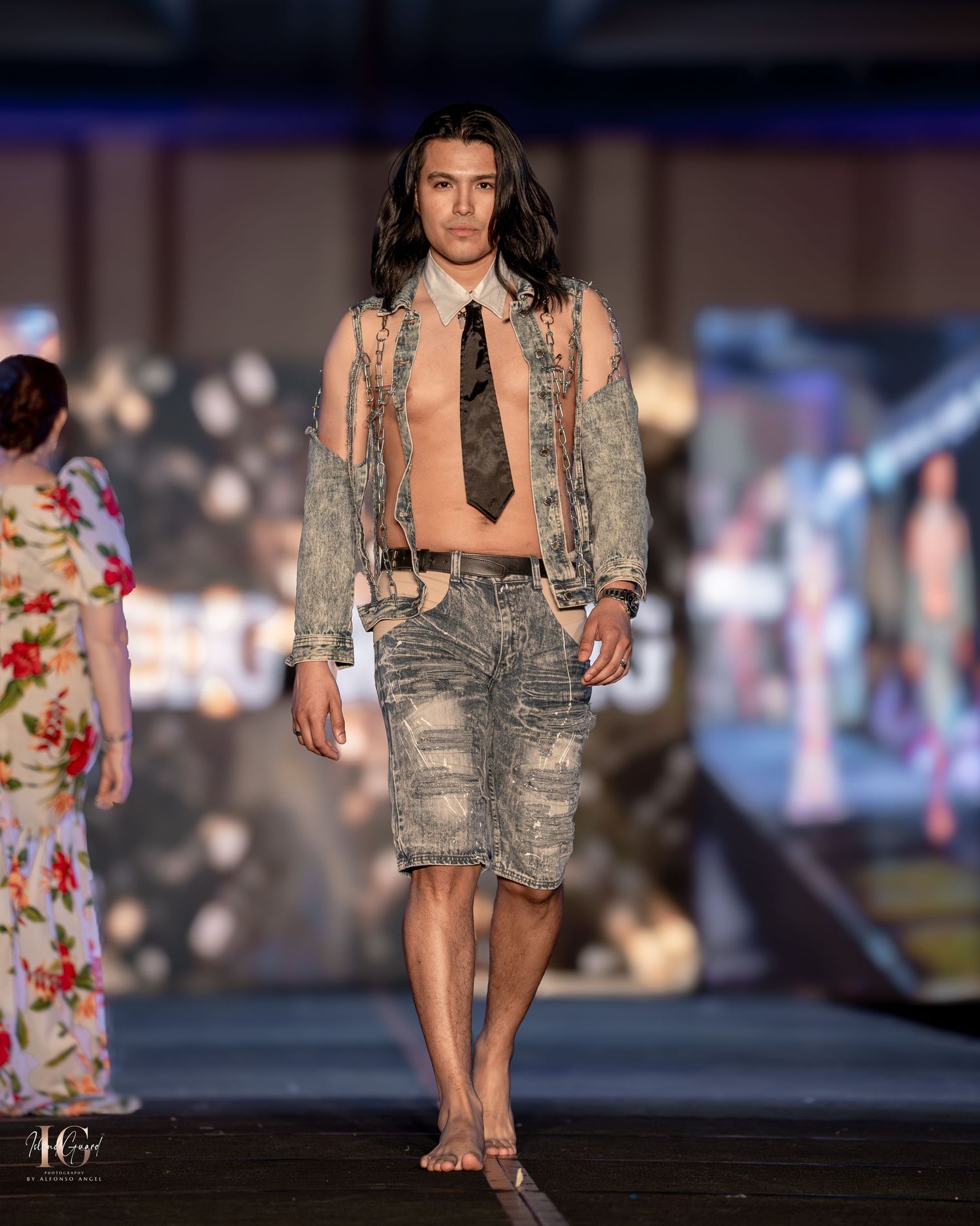 Model showcases distressed denim shorts with beige jacket and tie on fashion runway.