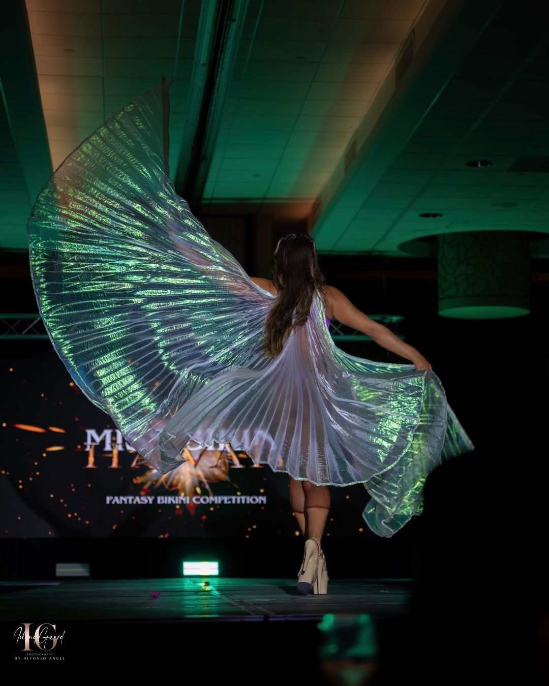 A dancer in a flowing white dress performs with illuminated LED wings glowing in turquoise light on a dark stage.