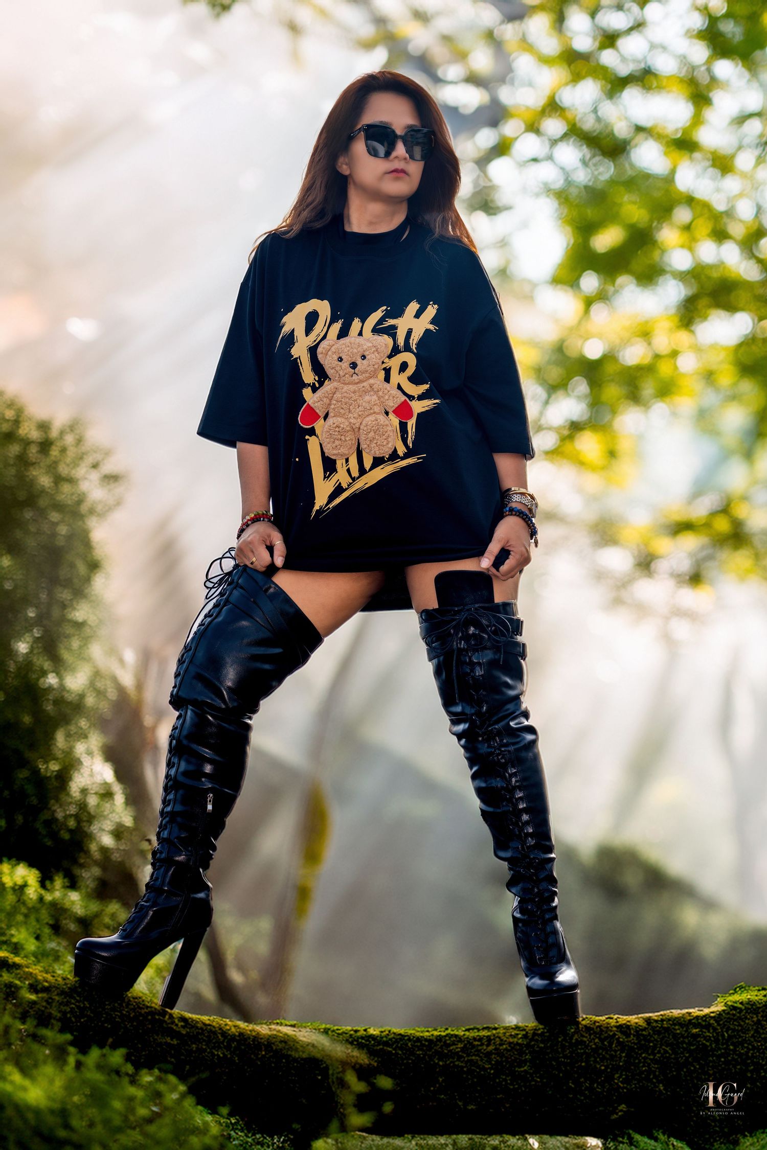 Person in black graphic t-shirt and boots posing against sunlit forest backdrop.