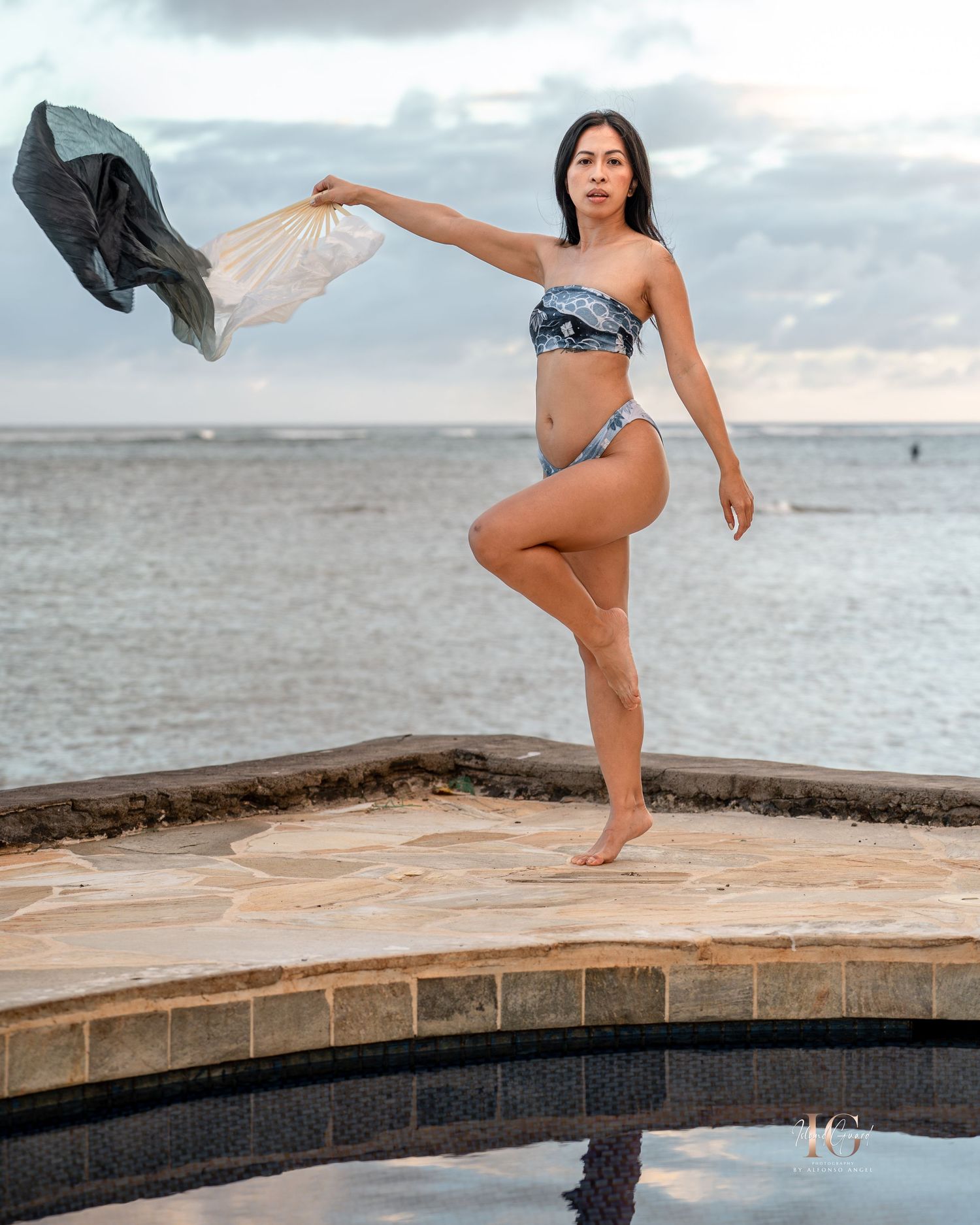 A person performs a graceful dance movement on a curved stone wall by the ocean at sunset.
