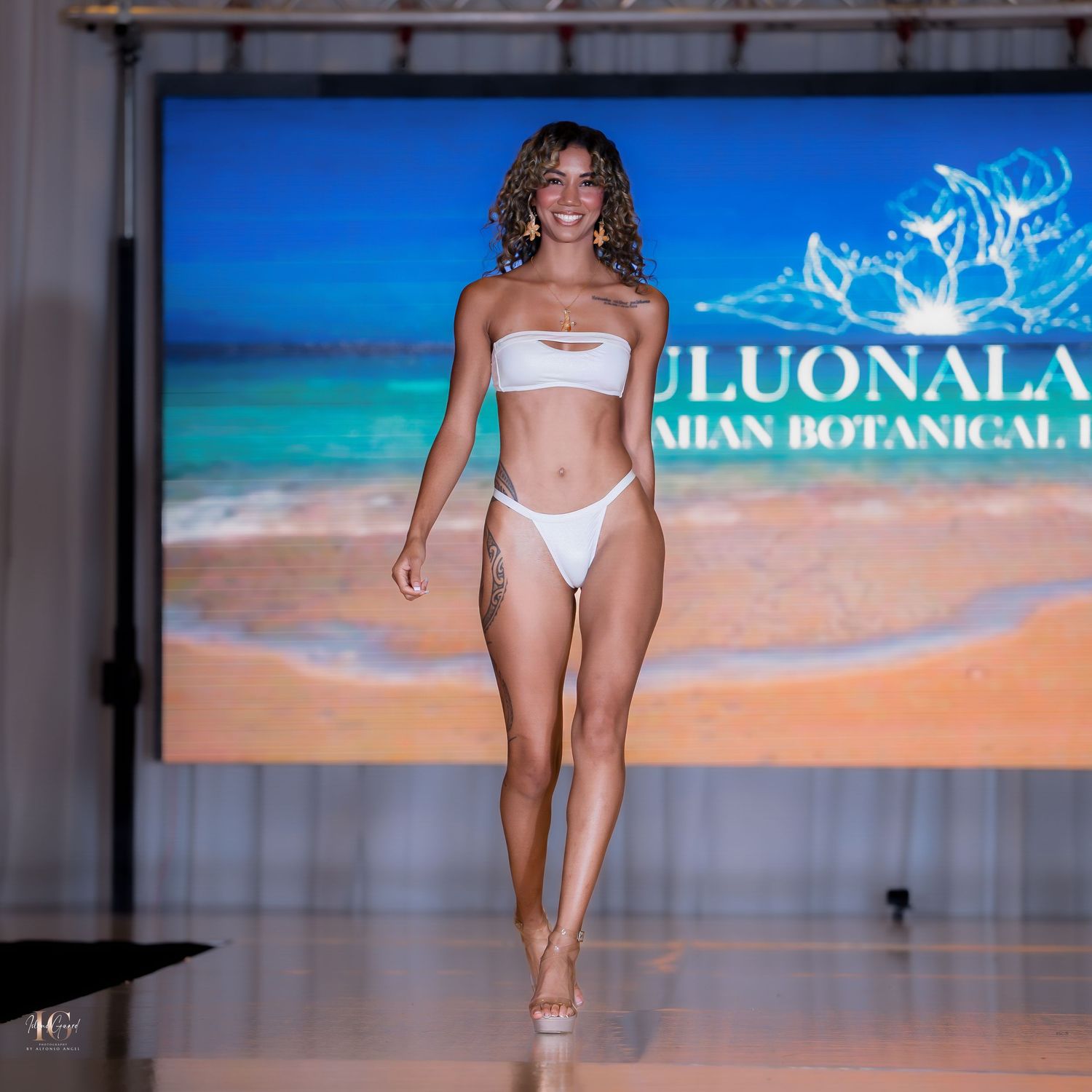 Models walk the runway in white swimwear during a beachwear fashion show with tropical backdrop.