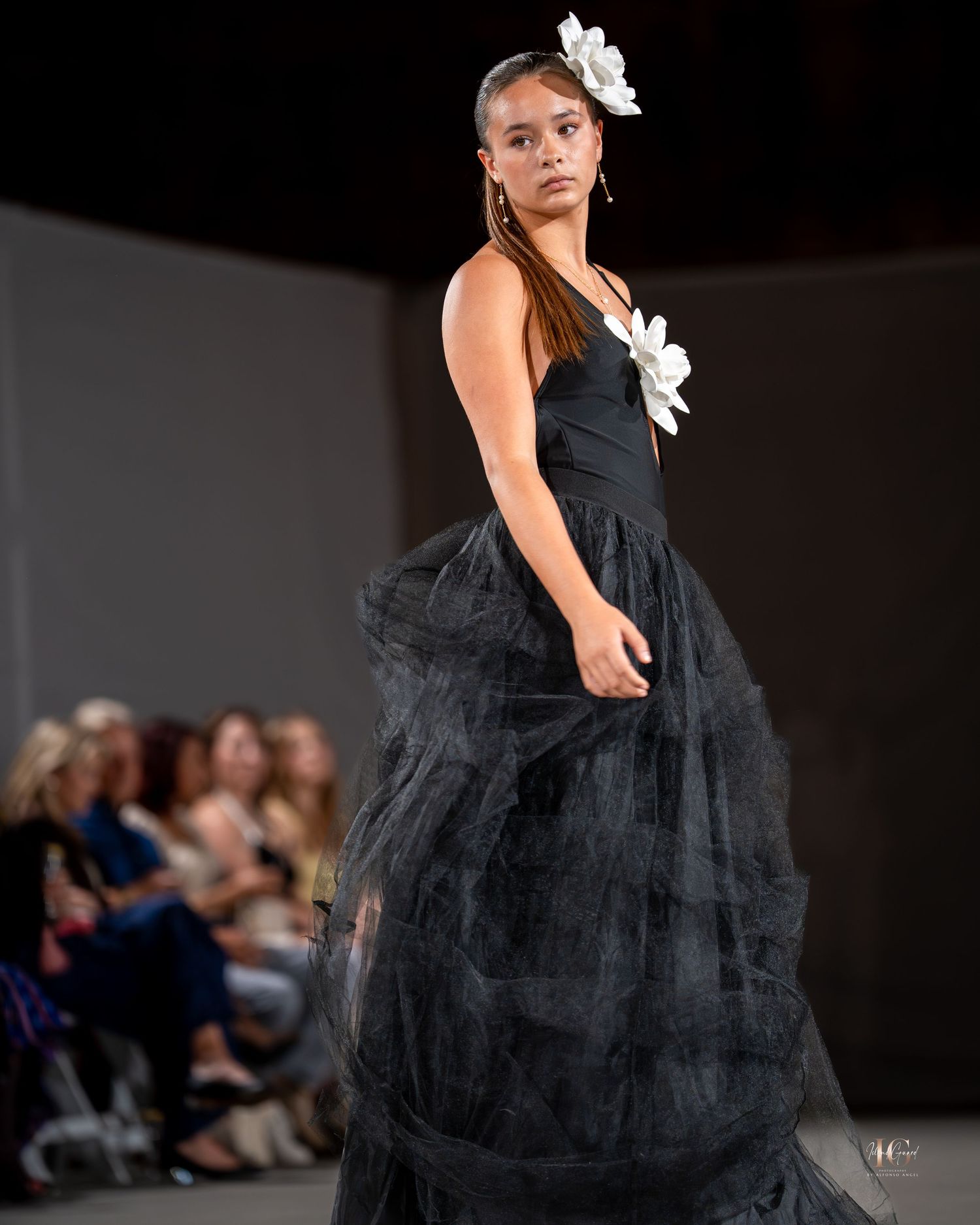 Model walks runway in dramatic black tulle gown with white floral accent during fashion show.
