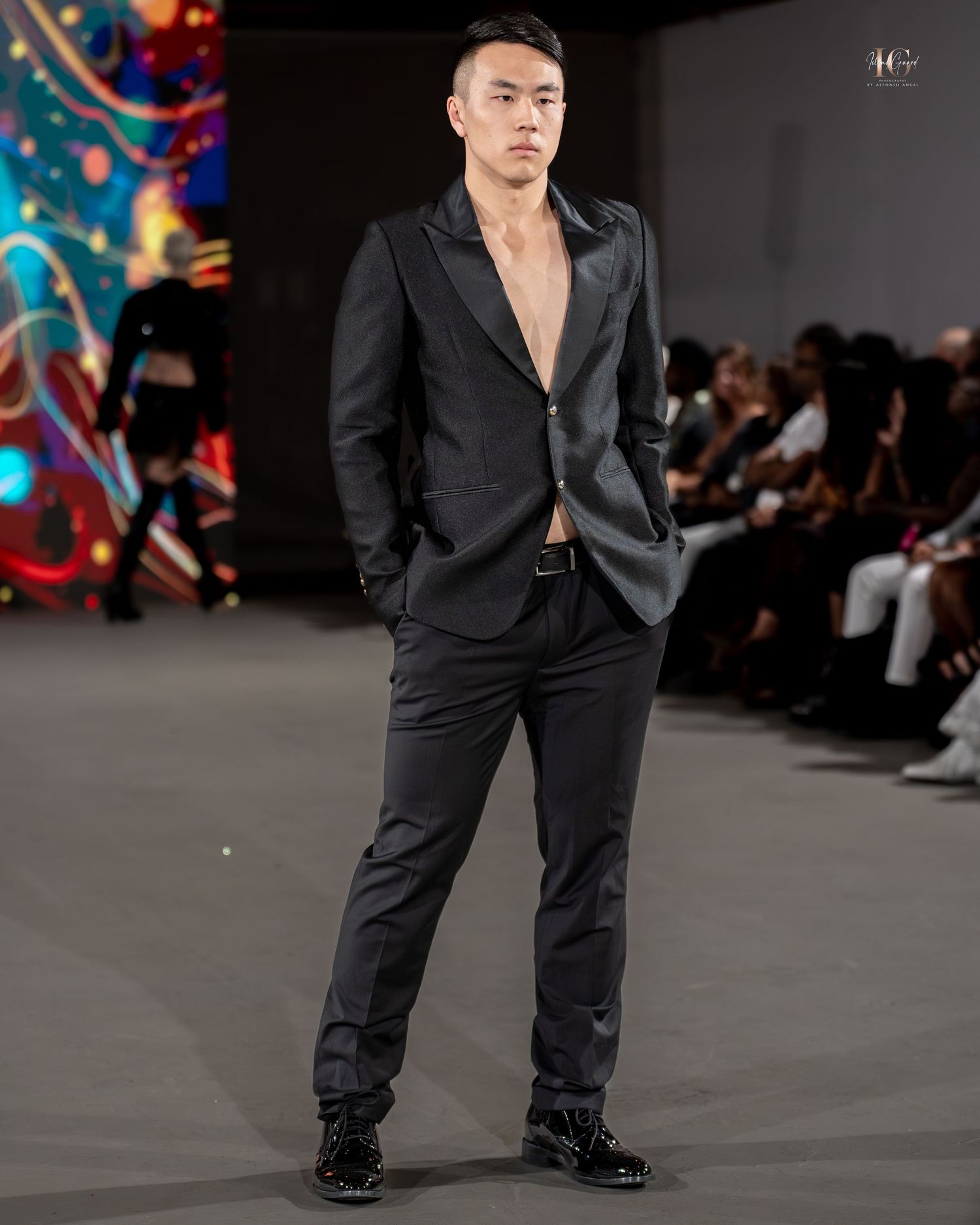 Model walks runway in all-black ensemble with loose-fitting shirt and pants during fashion show presentation.