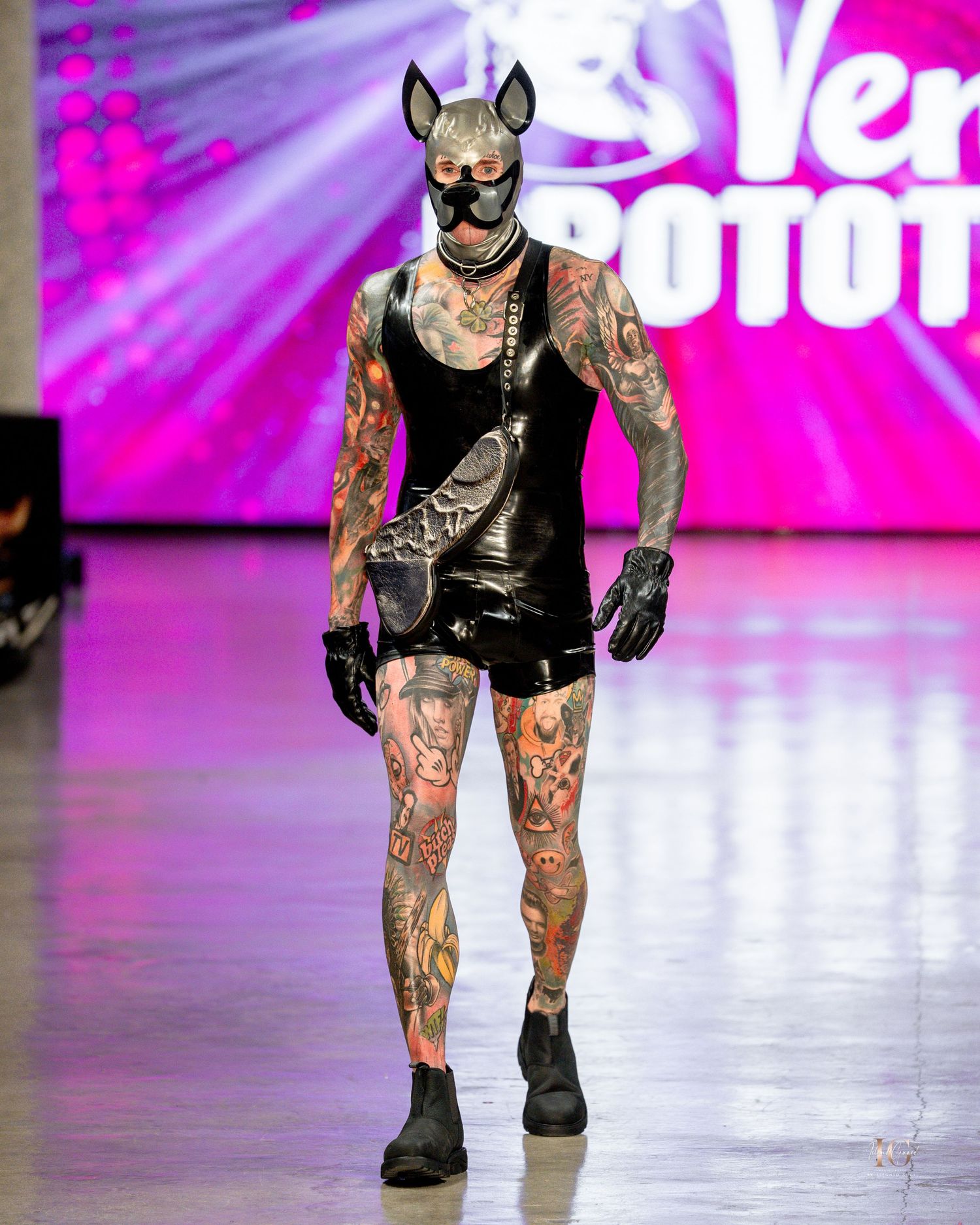 A performer in a black leather outfit with mouse ears and tattoos walks down a fashion runway with pink lighting.