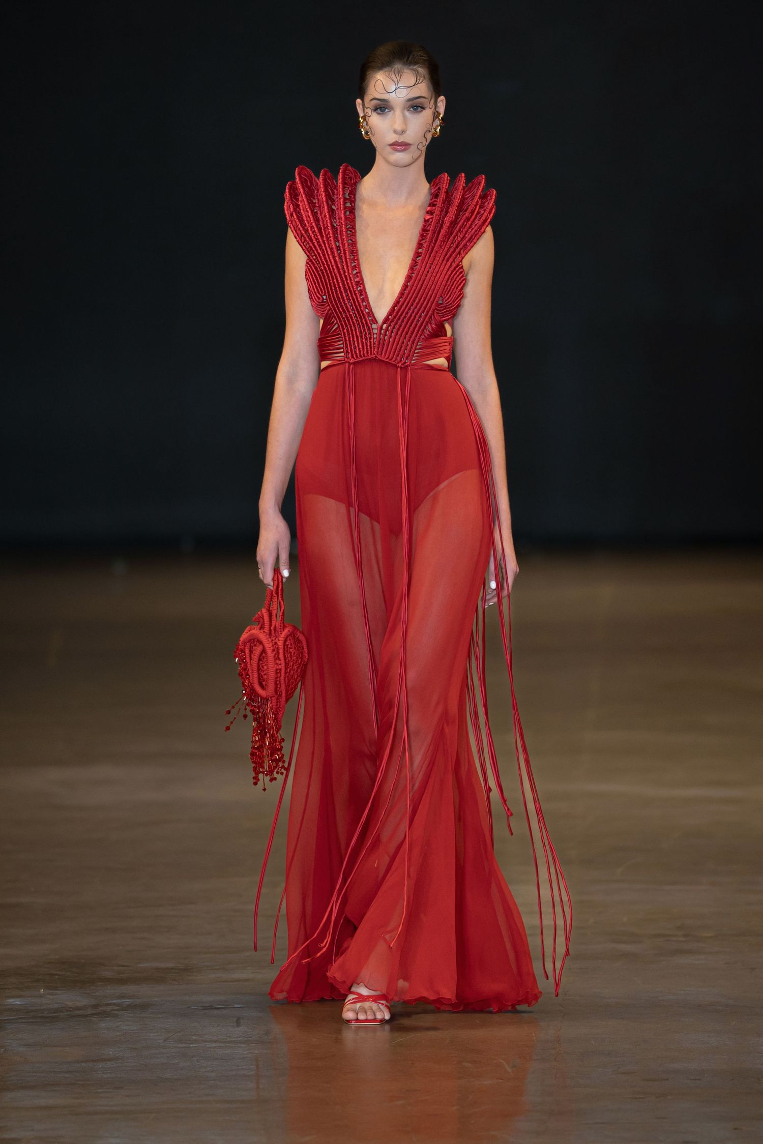 Model walks runway in flowing red sheer dress with ruffled details and matching accessories at fashion show.