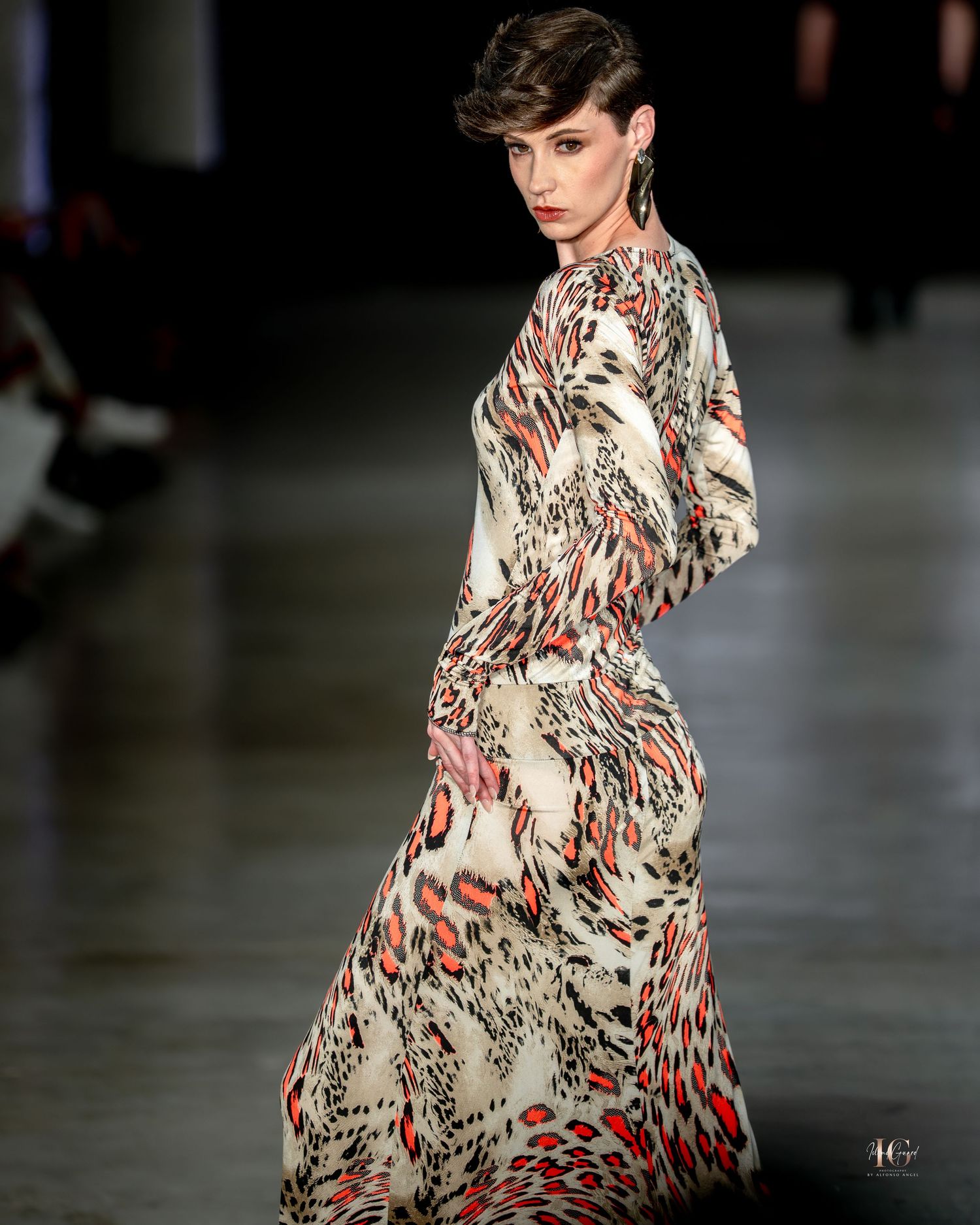 Model wearing a flowing abstract print dress in red, black and white patterns walks down a dark runway.