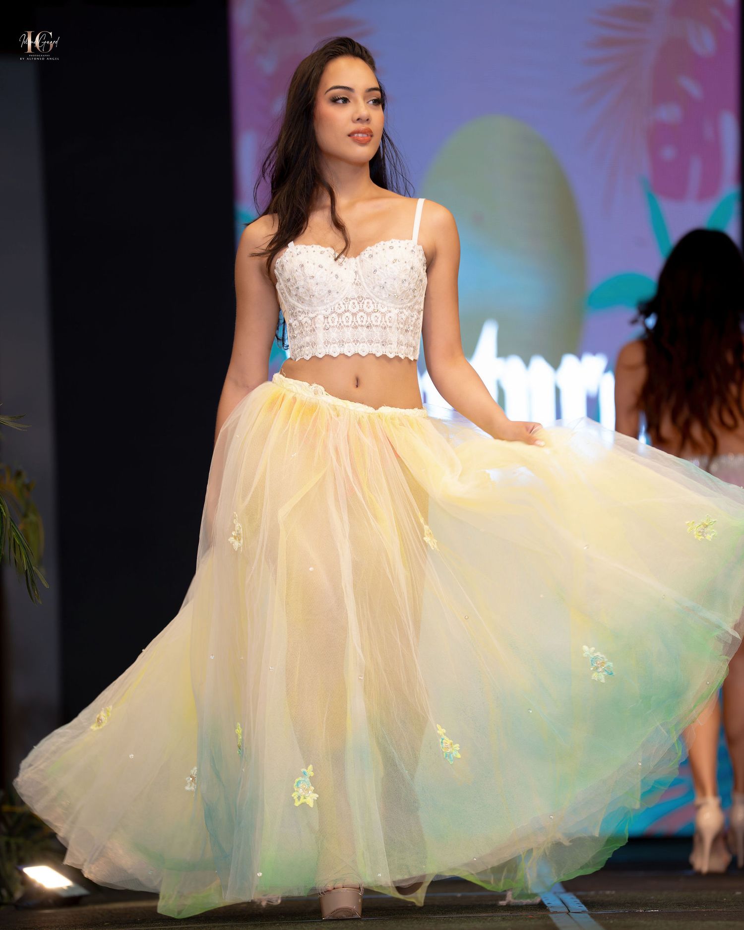 Model walks runway in white lace crop top and flowing yellow tulle skirt during fashion show.