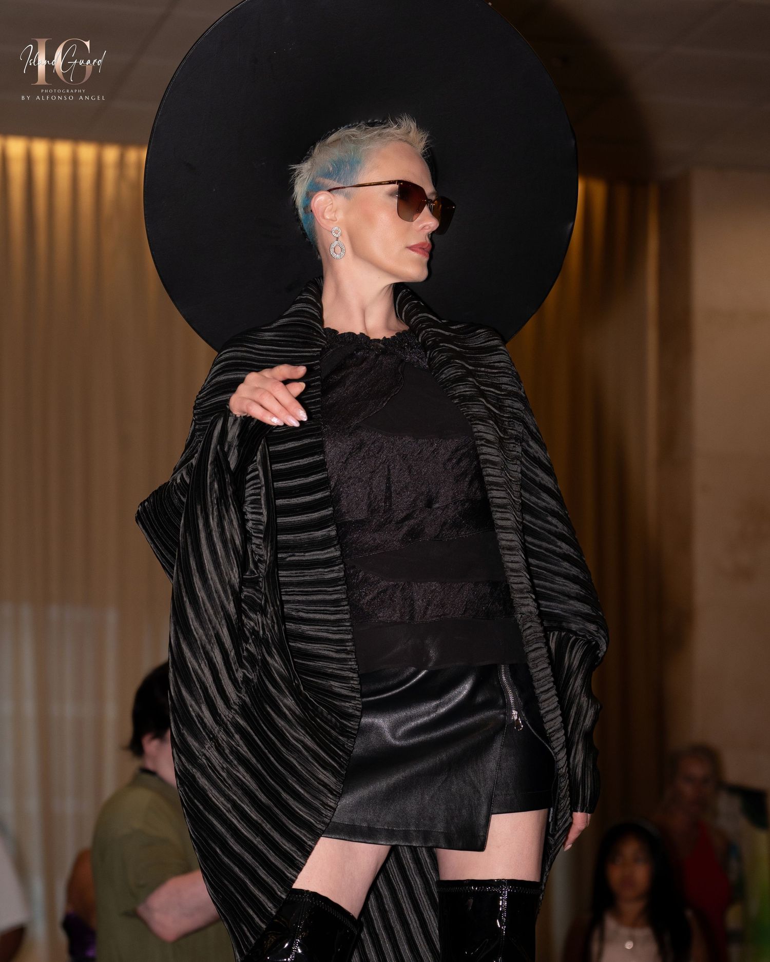 Model showcases black avant-garde fashion with dramatic striped cape and sleek silhouette on the runway.
