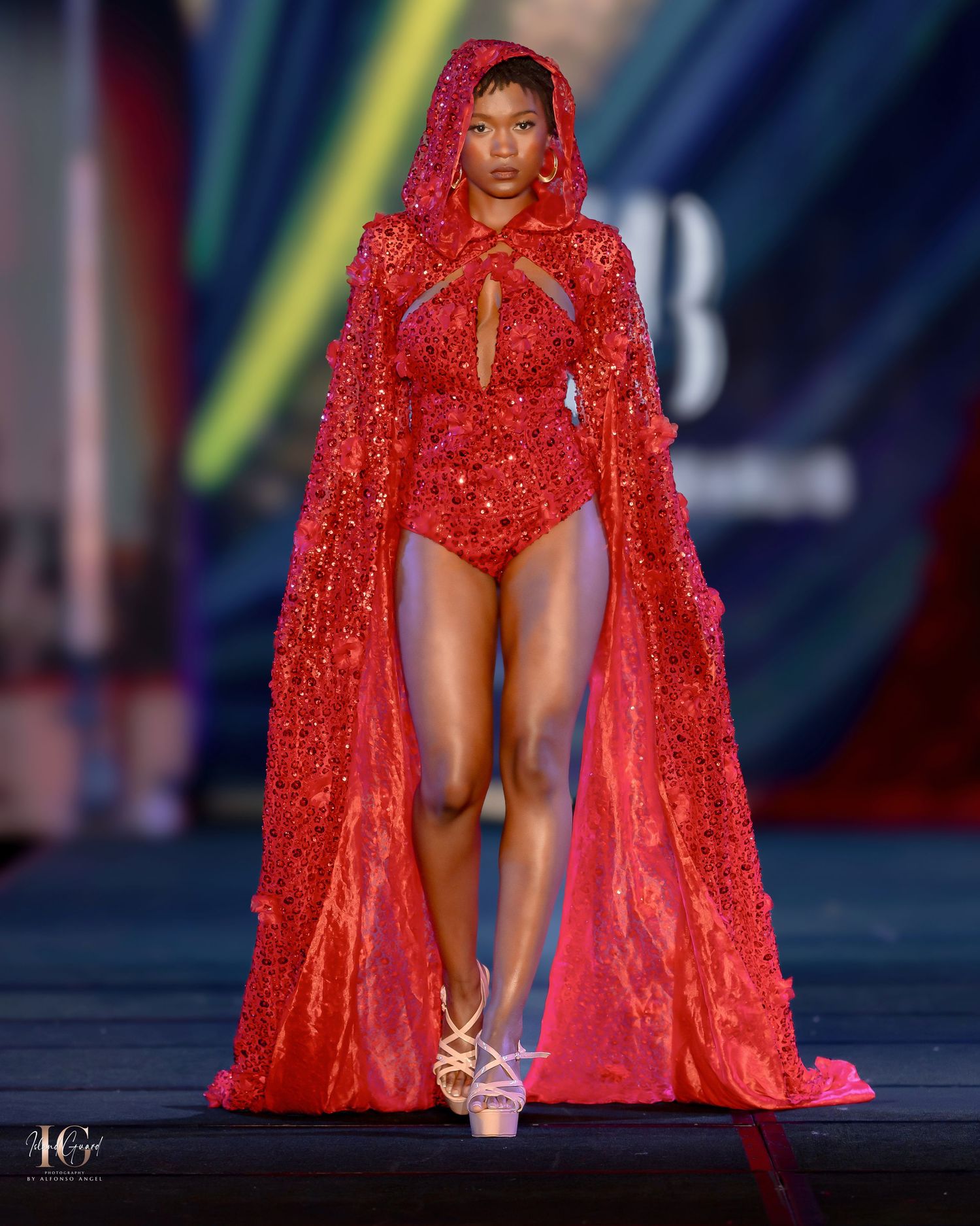 Dramatic red lace cape ensemble displayed on fashion runway with theatrical lighting.