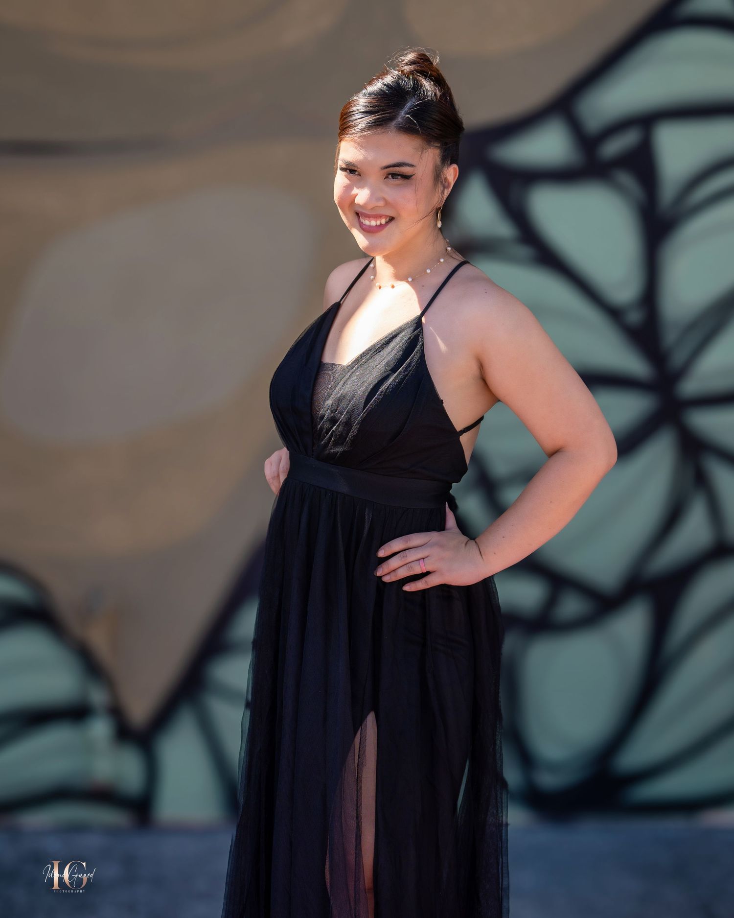 Model wearing flowing black dress poses against urban wall with artistic lighting.